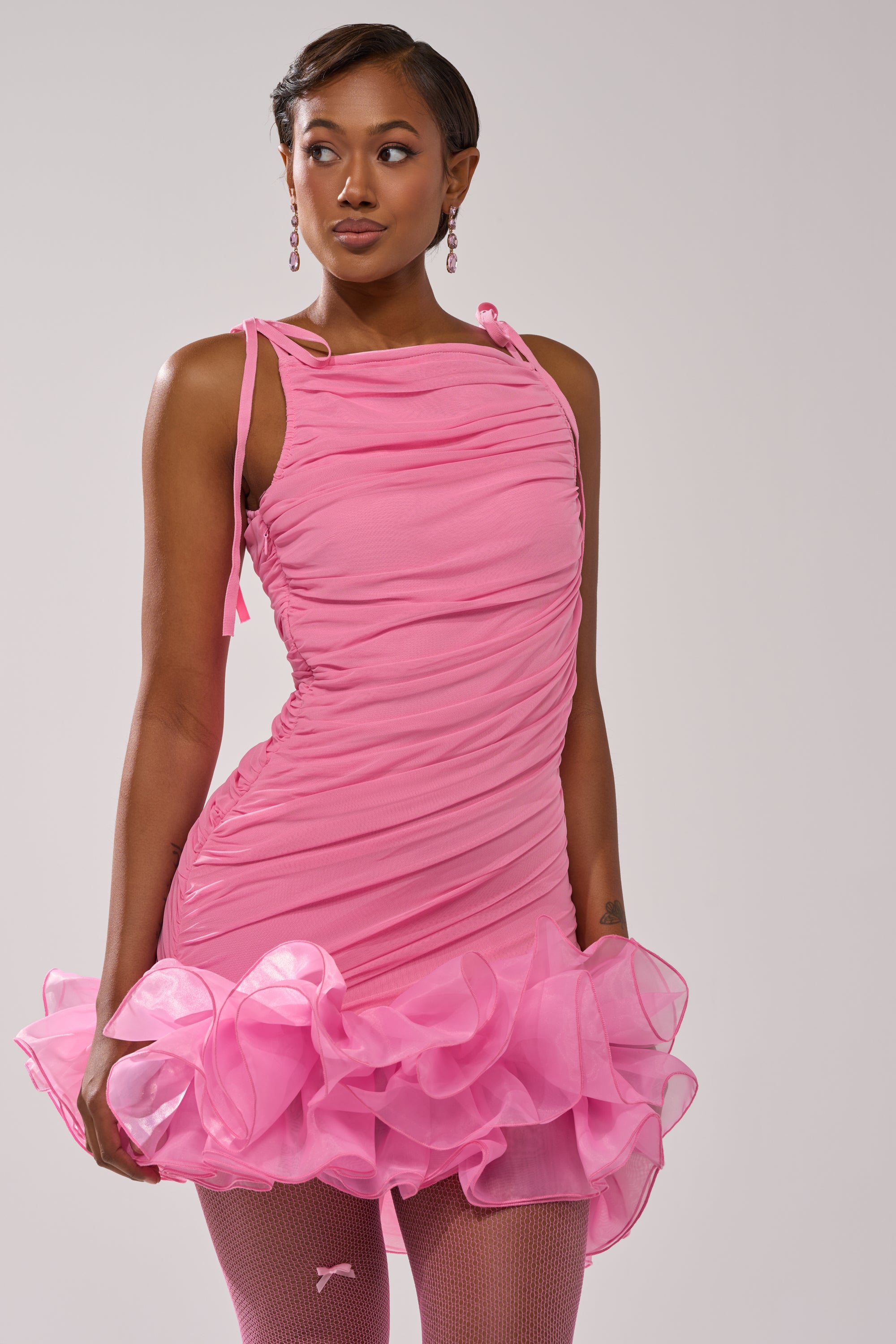 A woman in the PRINCESS PEPLUM MINI DRESS IN PINK—a fitted, ruched mesh mini dress with a ruffled hem—pairs it with pink earrings and fishnet tights, standing confidently against a plain background. 1