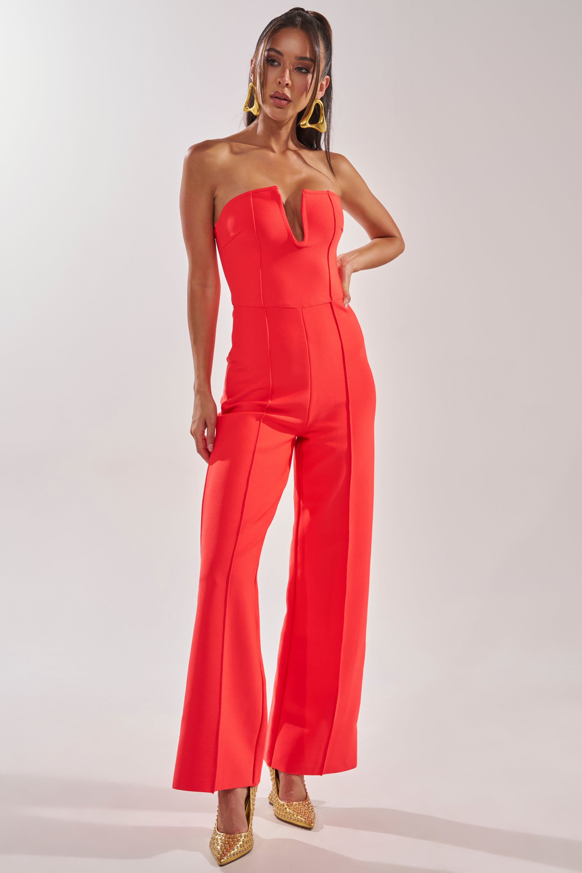 A woman stands against a plain light background in the BOSS LADY JUMPSUIT IN RED—strapless with a V-neckline and wide-leg pants—styled with gold hoop earrings and pointed-toe heels for a bold chic evening look. 0