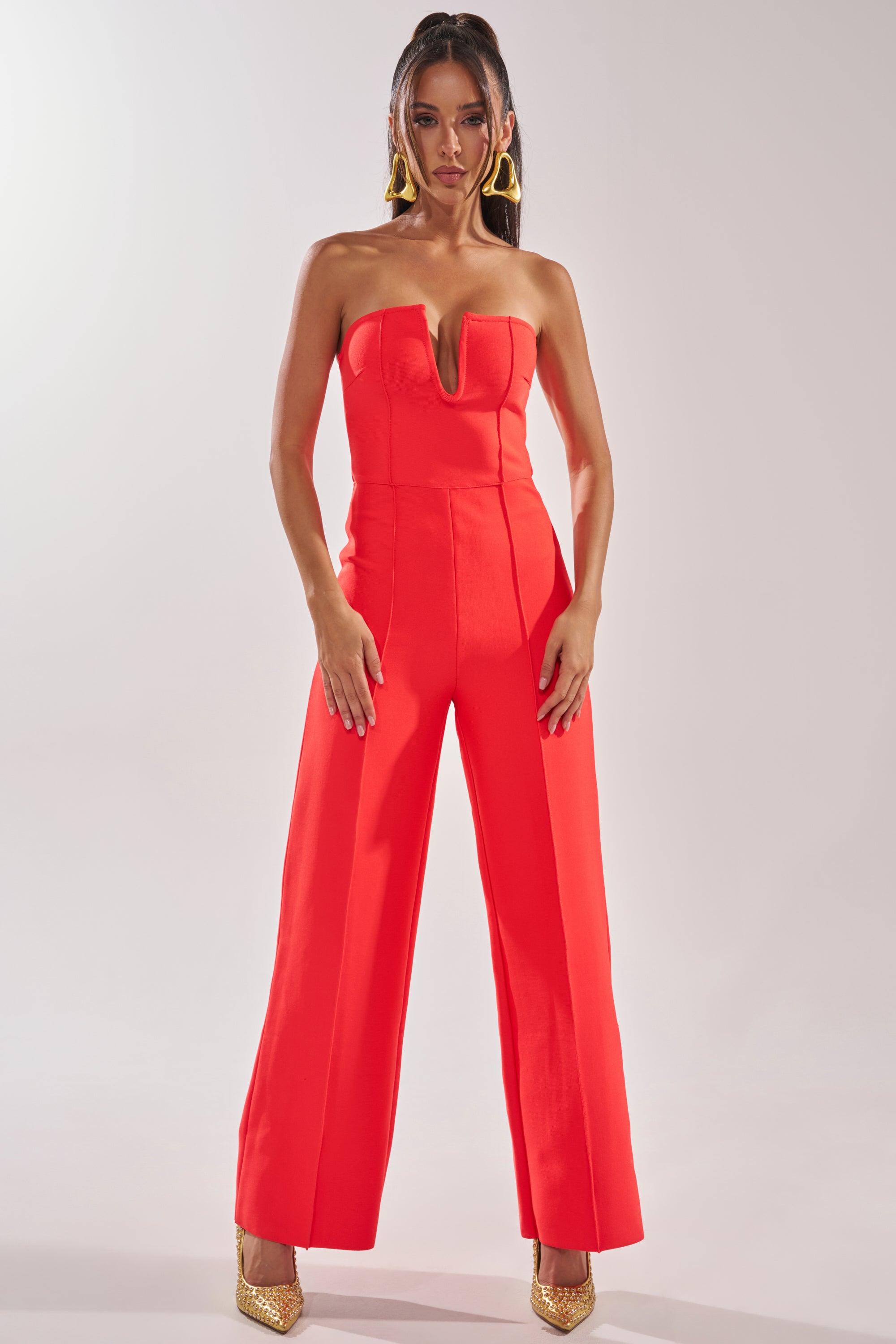 A woman models the BOSS LADY JUMPSUIT IN RED, featuring a strapless design, wide-leg pants, and a sharp V-cut neckline. She completes the look with long hair, bold gold earrings, and gold heels against a plain light background. 1