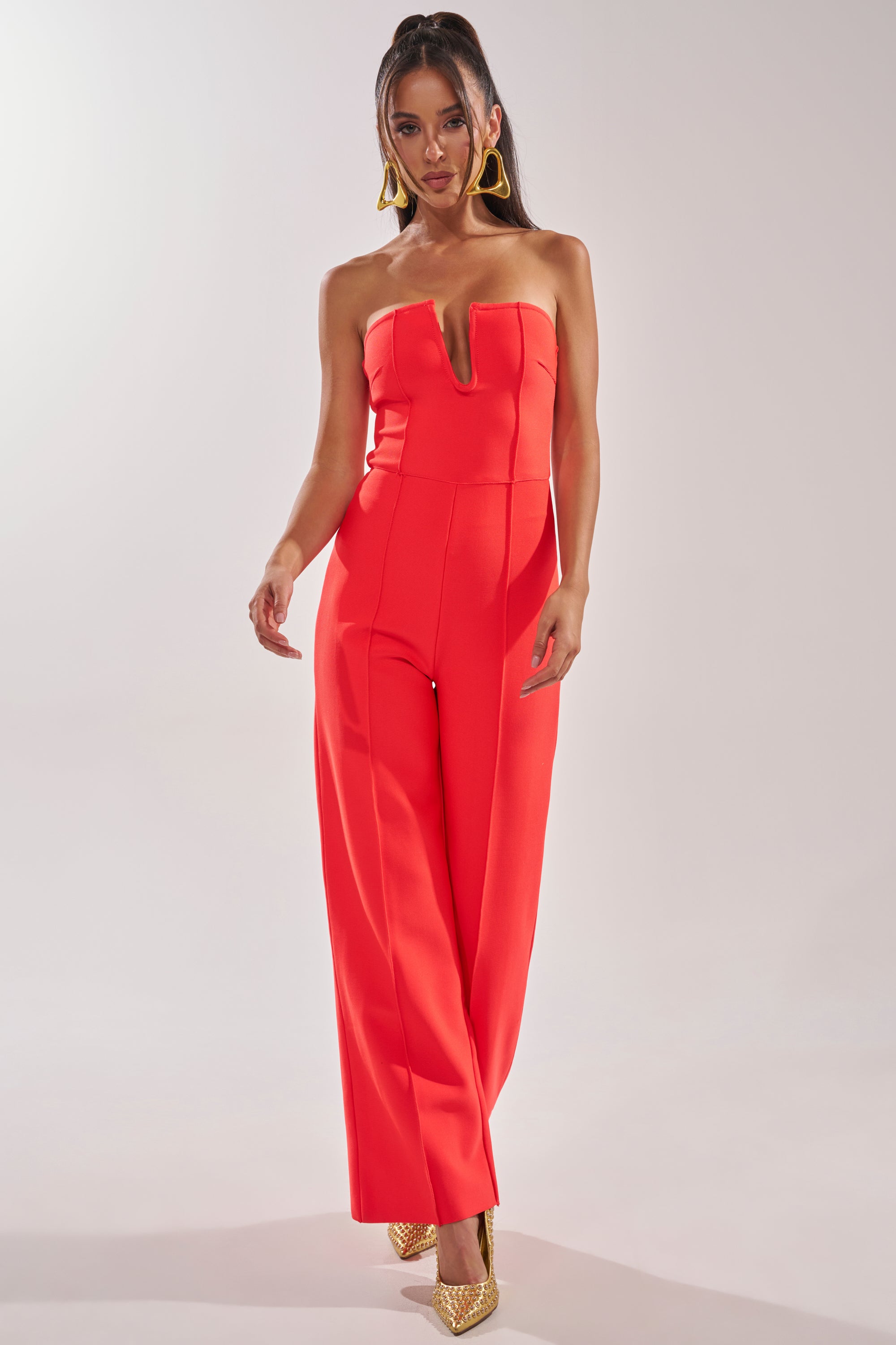 A woman models the BOSS LADY JUMPSUIT IN RED, a strapless wide-leg jumpsuit with a deep V neckline—ideal for formal evenings. She pairs it with statement gold earrings and heels, standing against a simple light background. 2