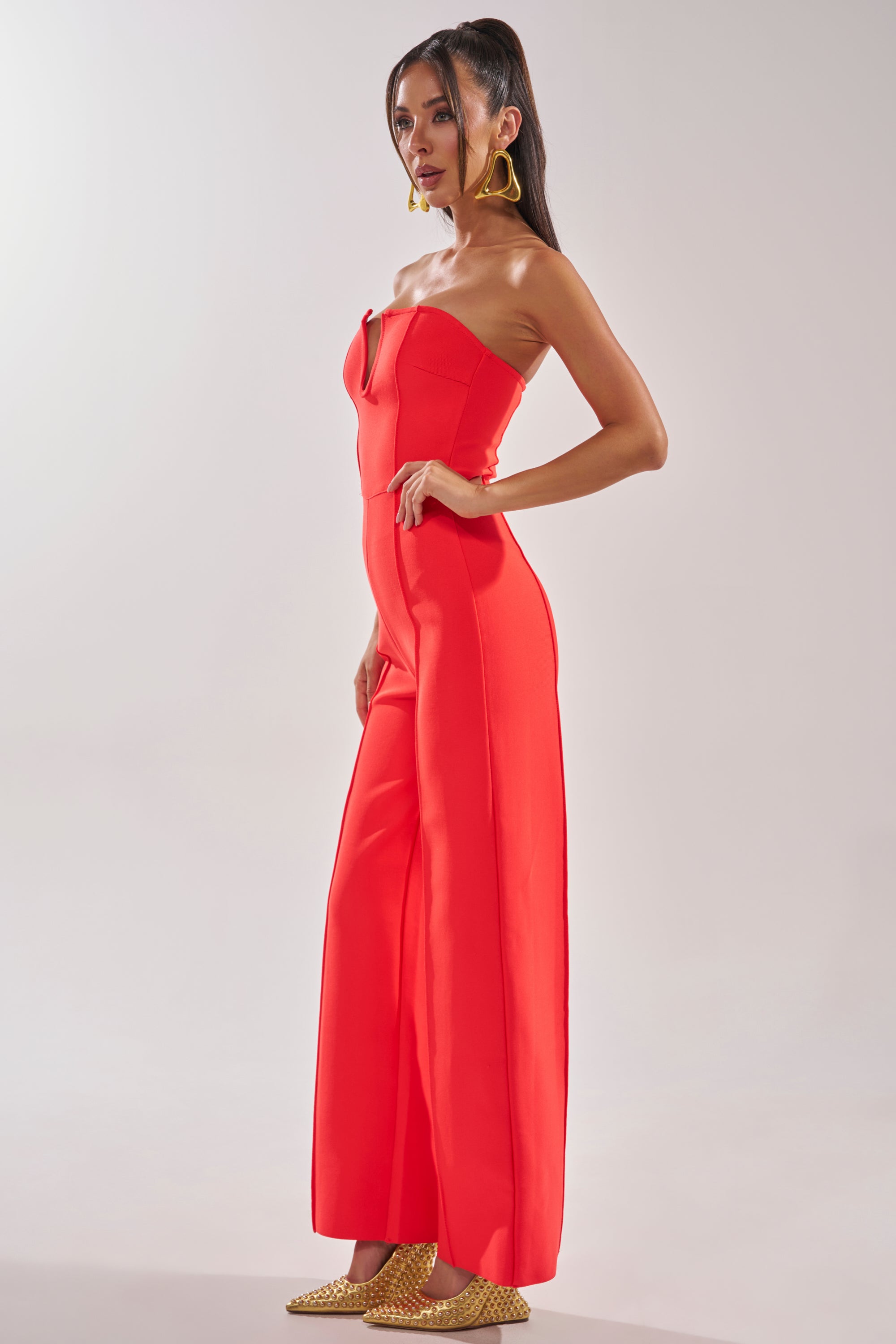 A woman stands in profile wearing the BOSS LADY JUMPSUIT IN RED with wide legs, gold pointed-toe heels, and large gold hoop earrings, embodying chic formal evening style against a plain light background. 3