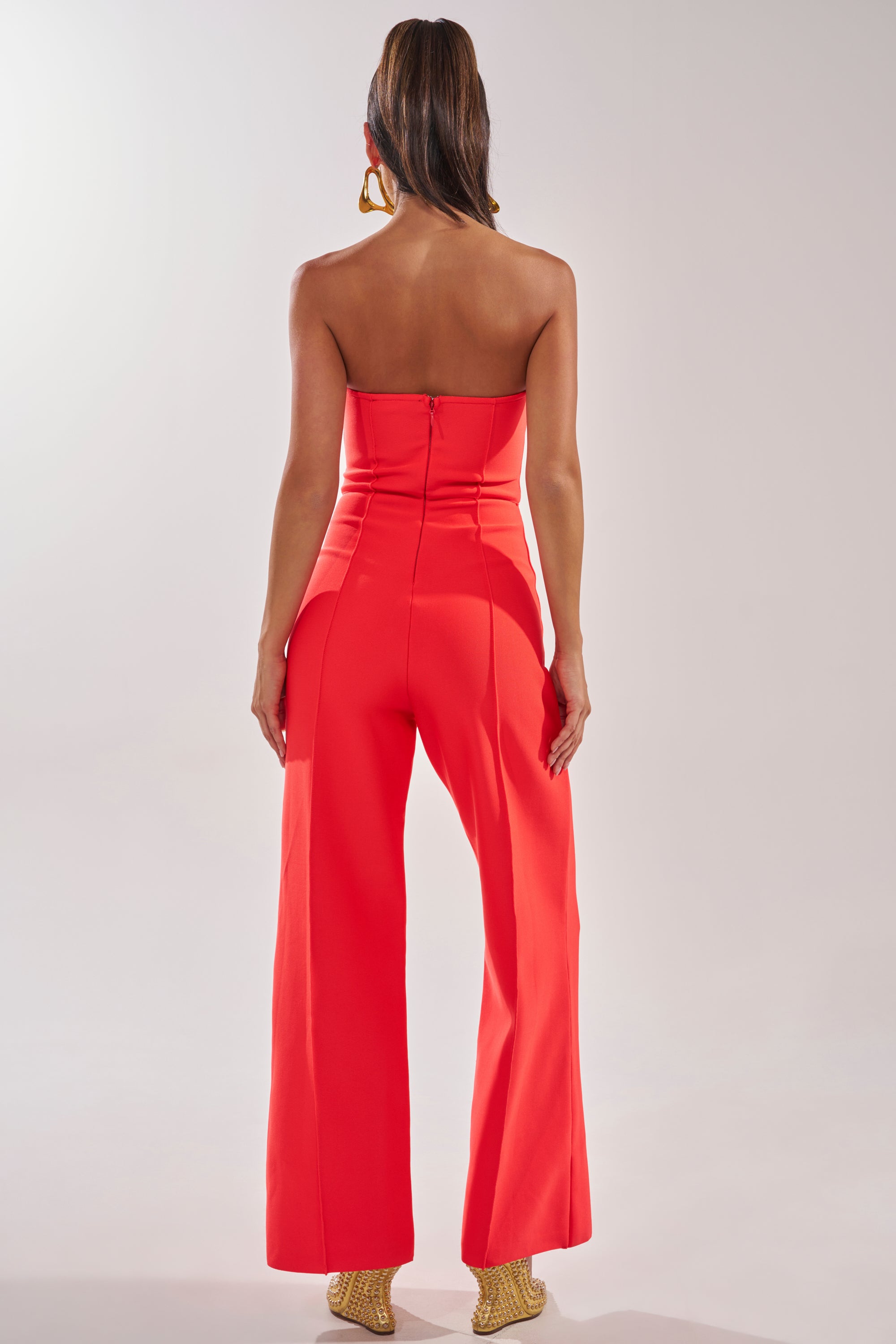 Woman stands with her back to the camera in the BOSS LADY JUMPSUIT IN RED, a strapless, wide-leg style paired with gold heels and hoop earrings—an effortlessly chic choice for formal evening wear. 4