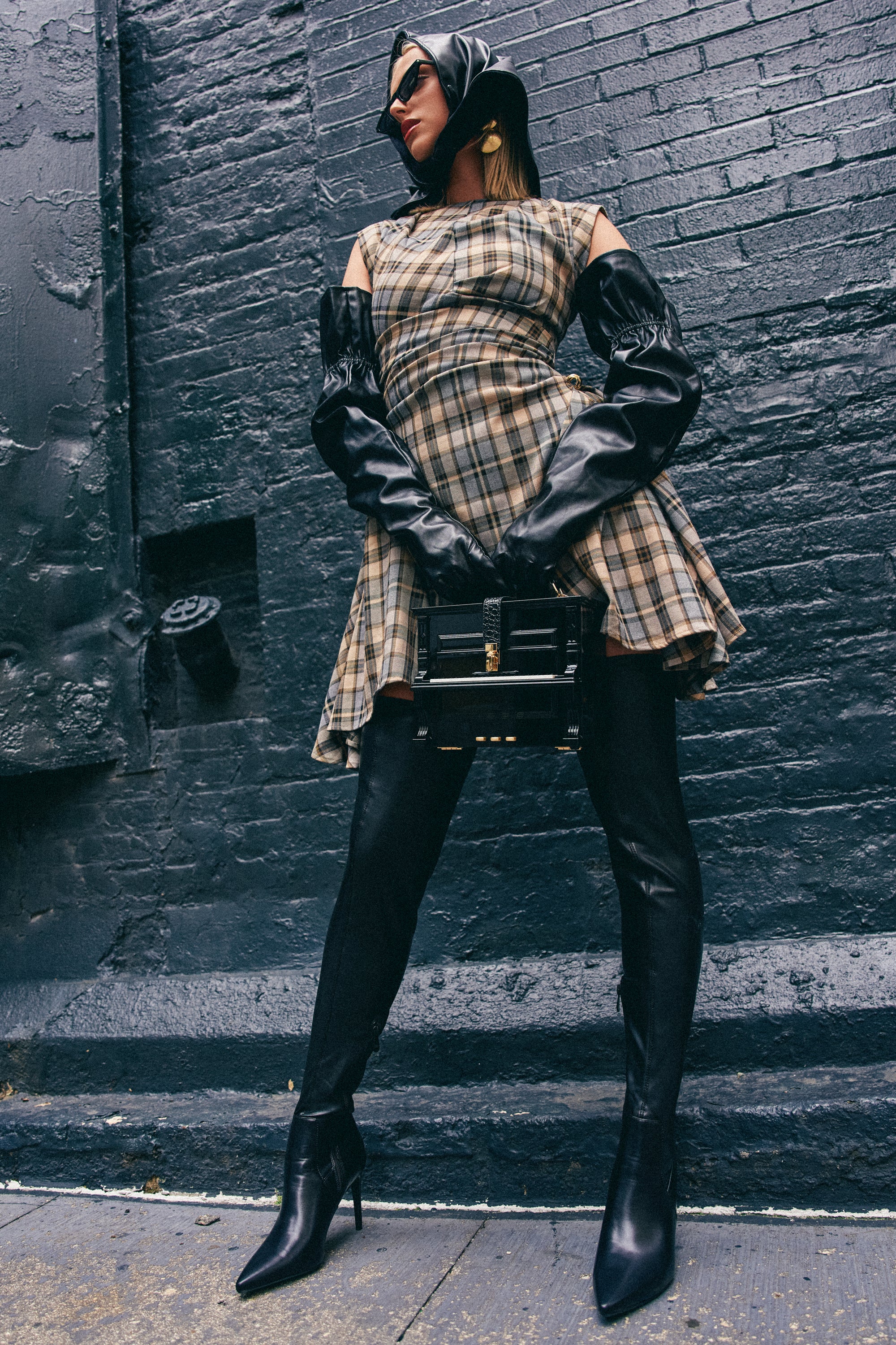 Wearing the CARRY ME HOME MINI DRESS IN PLAID, along with long black gloves, black heeled boots, a headscarf, and holding a black handbag, a person stands confidently against a dark brick wall.