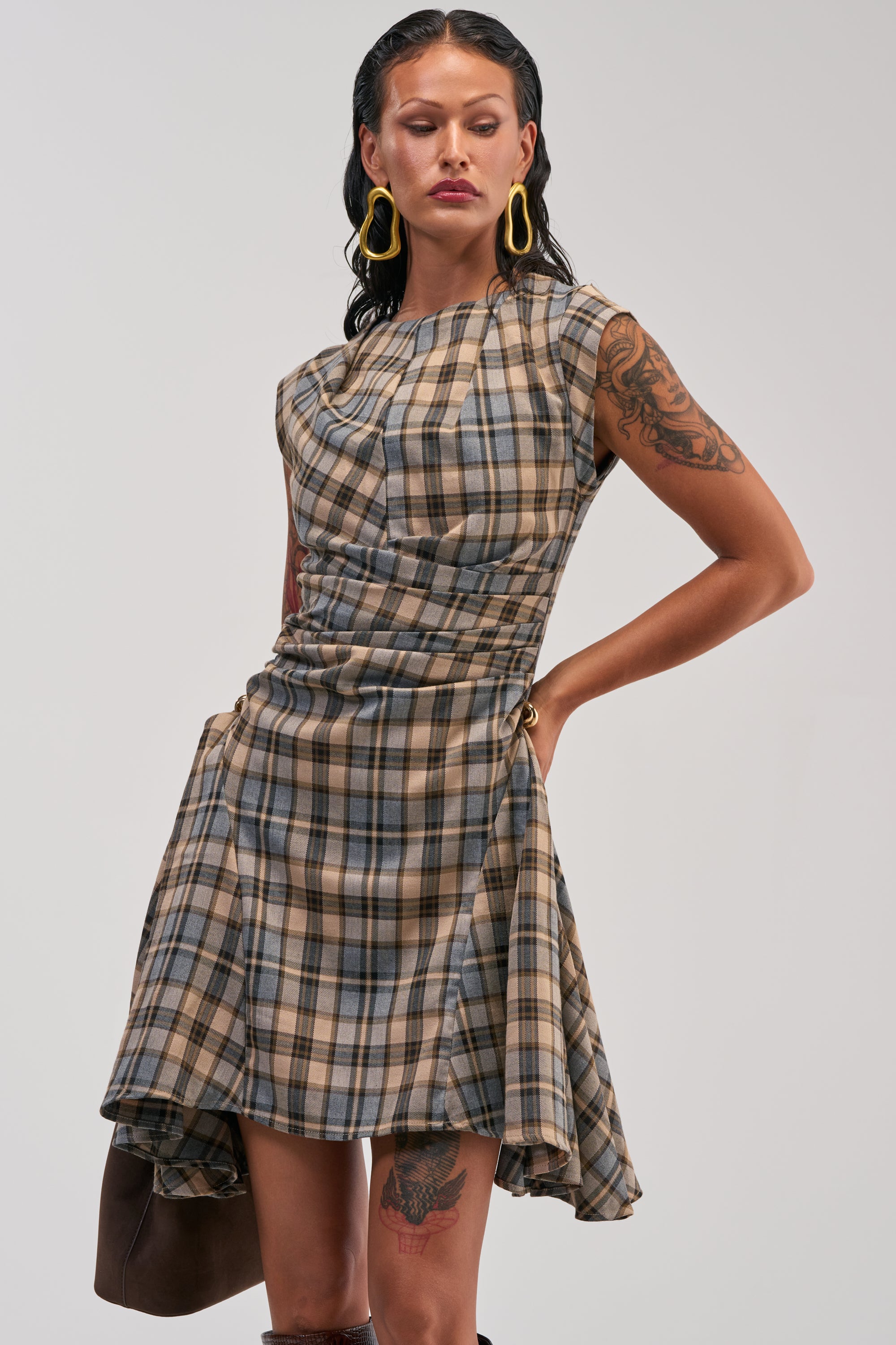 A woman showcases the CARRY ME HOME MINI DRESS IN PLAID—a sleeveless, asymmetrical mini dress featuring brown and blue plaid with ruched detailing—styled with yellow earrings and visible tattoos as she poses with one hand on her hip against a plain background.
