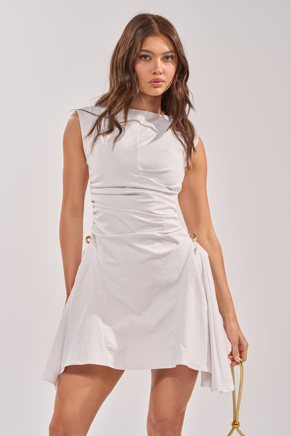 A woman with long brown hair wears the CARRY ME HOME MINI DRESS—a white, sleeveless ruched mini dress with gathered side details and an asymmetrical cut. She holds a gold accessory in her right hand against a light background. 0