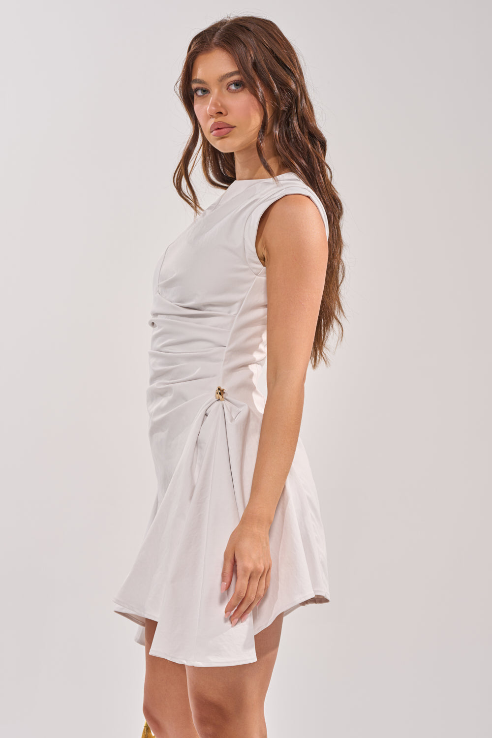 A woman with long brown hair wears the CARRY ME HOME MINI DRESS, a white, sleeveless, ruched mini dress with gold accents. She stands sideways against a plain light background, facing the camera in this bridal-inspired look. 2