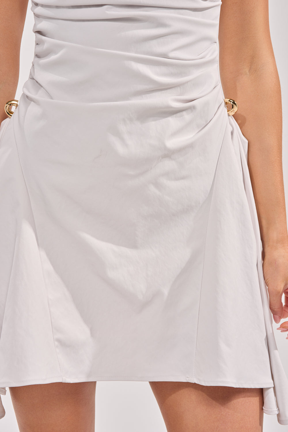 A close-up of a person wearing the CARRY ME HOME MINI DRESS in light gray, featuring a ruched fit-and-flare design with gathered fabric and gold ring accents at the hips. Only their lower torso and upper legs are visible. 5