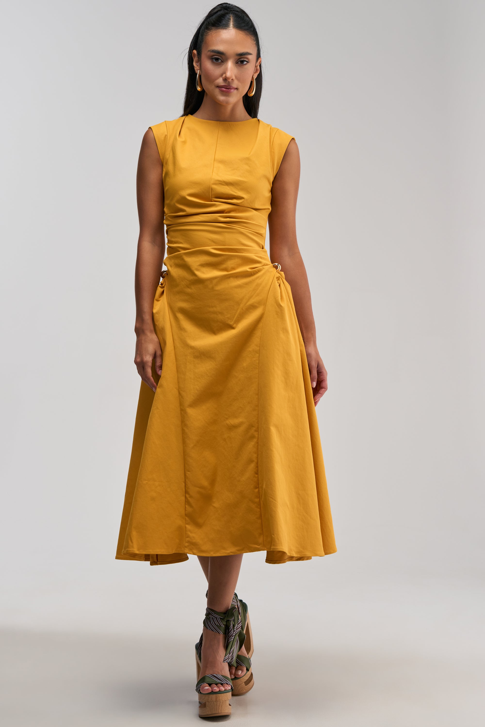 A woman wearing the IT’S MY TIME MIDI DRESS IN MUSTARD, a sleeveless midi dress featuring a ruched waist and gold ring details, paired with green strappy cork wedge sandals and gold hoop earrings—an effortlessly stylish business casual look perfect for any work event.
