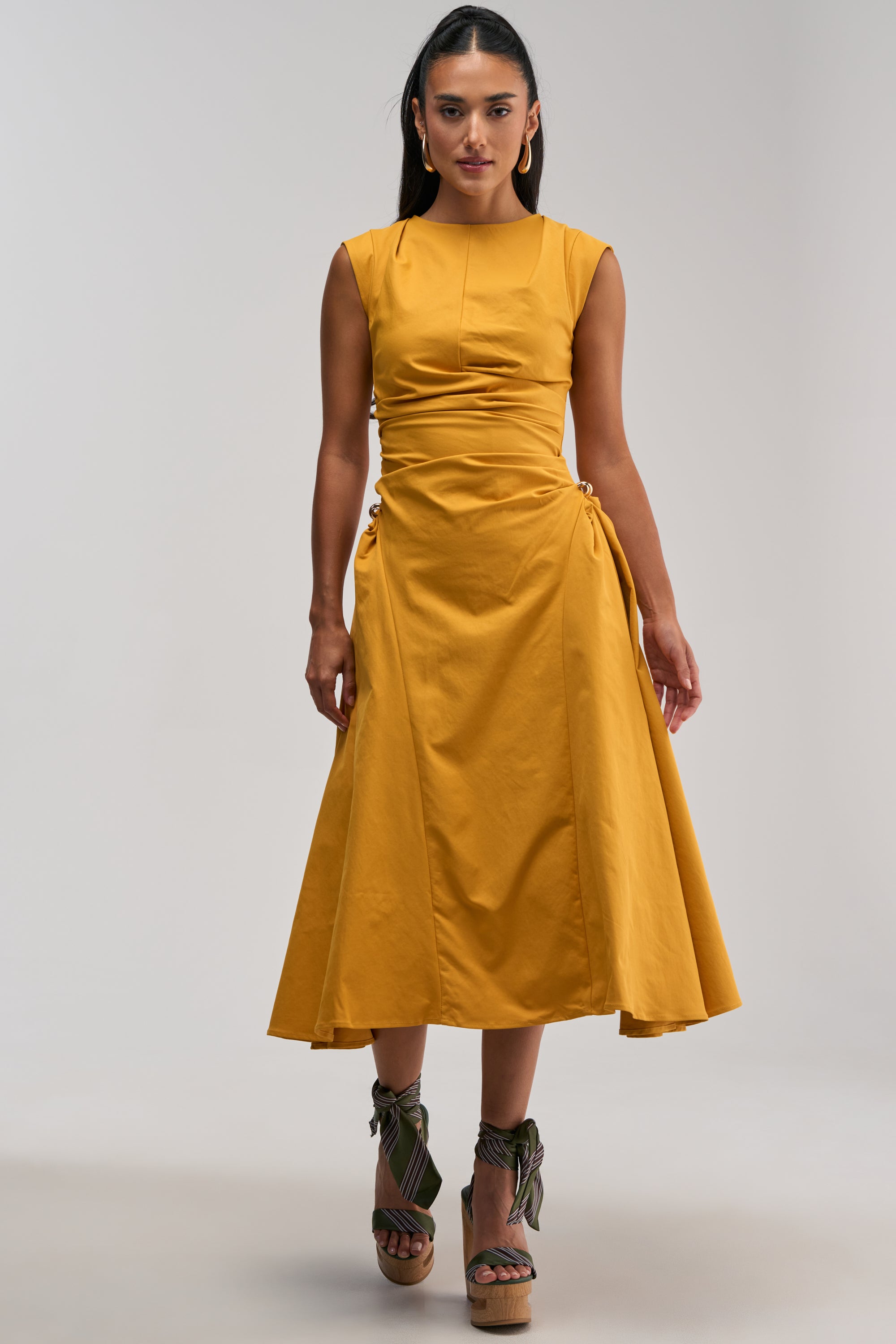 A woman wearing the IT’S MY TIME MIDI DRESS IN MUSTARD—featuring waist ruching and tied details at the sides—pairs it with open-toe wedge sandals with ankle ties. Perfect for business casual occasions or work events, she stands against a plain, light background.