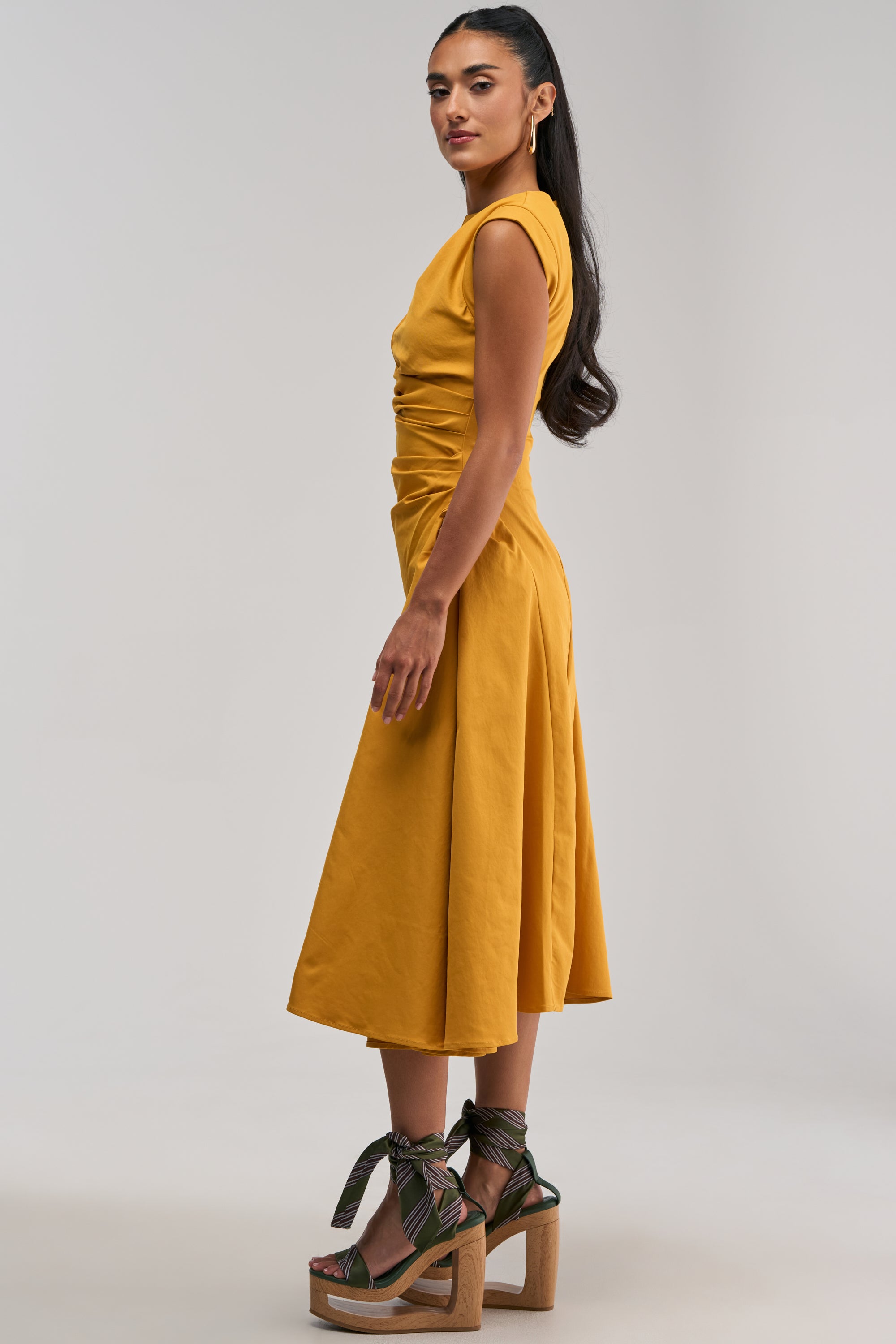 Wearing the IT’S MY TIME MIDI DRESS IN MUSTARD—a stylish midi dress featuring cap sleeves and flattering side ruching—a woman stands sideways in platform heels with green ankle ties against a plain light background, creating the perfect statement for a chic business casual dress code.