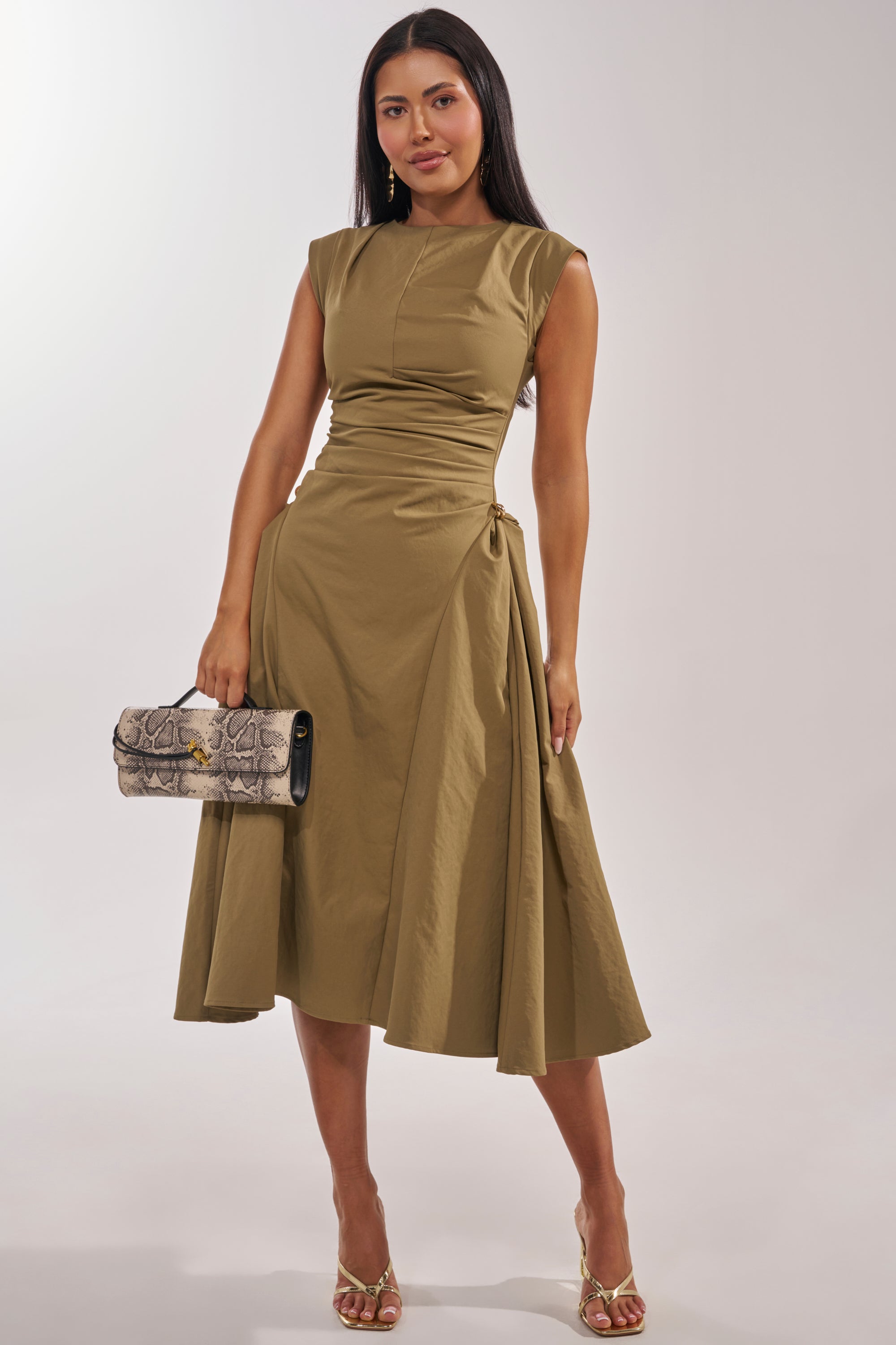 A woman wears the IT'S MY TIME MIDI DRESS IN OLIVE, featuring a high neck and ruched details, paired with gold heels and a gray snakeskin clutch as she smiles at the camera against a light background. 0