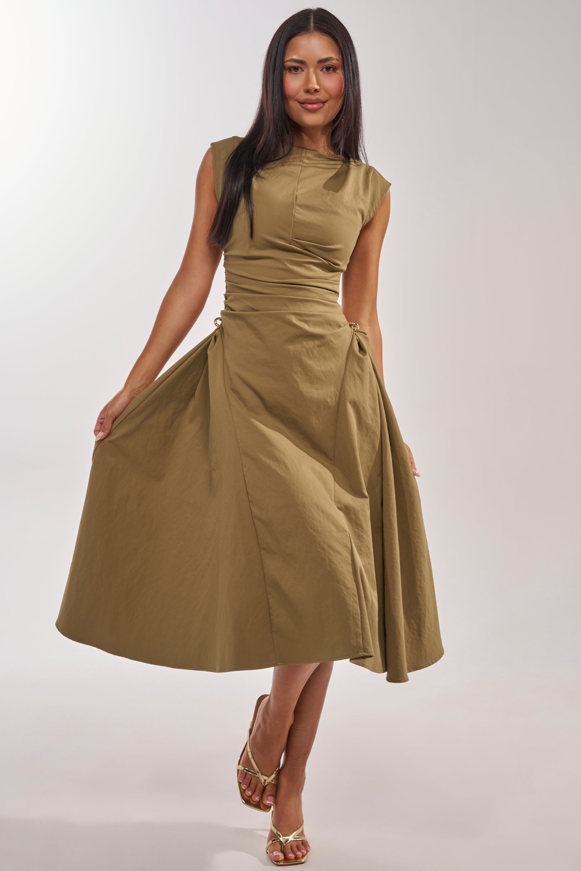 A woman stands against a light background, smiling as she holds out the skirt of the IT'S MY TIME MIDI DRESS IN OLIVE, featuring a high neck, ruched sides with ties, and gold accents. She pairs it with open-toe sandals. 1