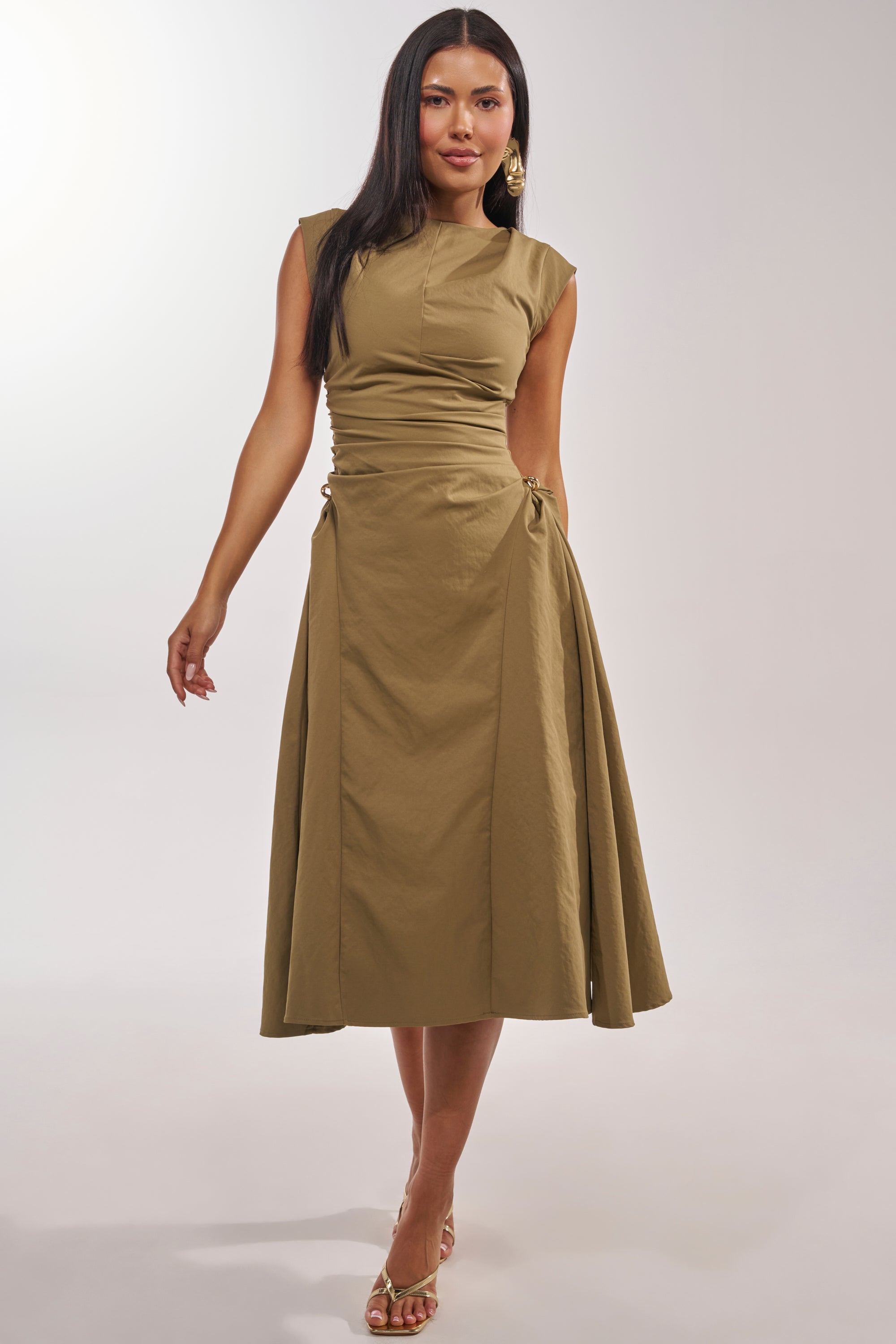 A woman models the IT'S MY TIME MIDI DRESS IN OLIVE, a sleeveless midi dress with a fitted bodice and sculptural silhouette, styled with gold earrings and heeled sandals against a plain light background. 2