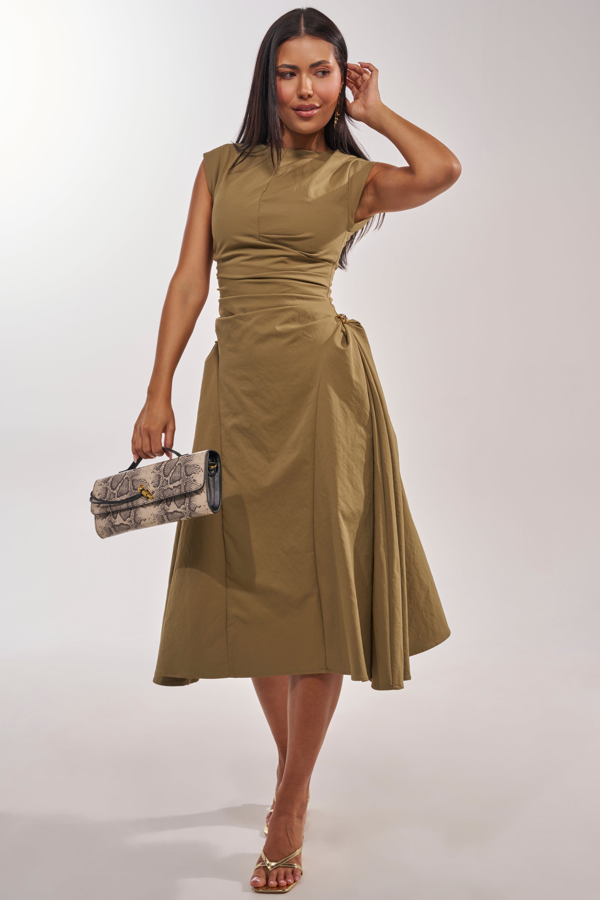 A woman wears the IT'S MY TIME MIDI DRESS IN OLIVE, featuring cap sleeves and gold accents. She pairs it with strappy gold heels and a snakeskin clutch, posing with one hand on her head and her long hair straightened. 3
