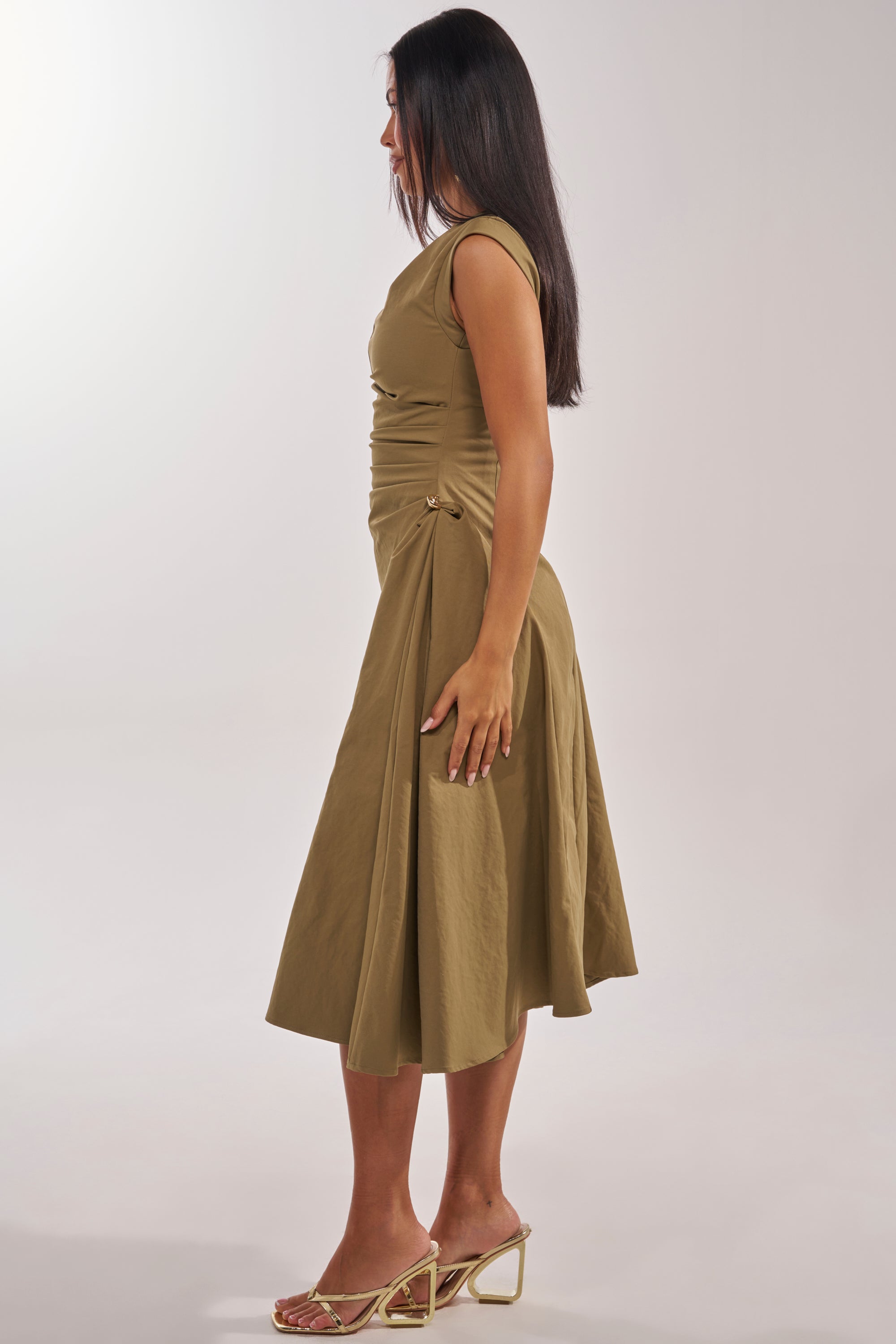 A woman with long dark hair stands in profile against a plain light background, wearing the IT'S MY TIME MIDI DRESS IN OLIVE with a high neck, ruched details, sculptural silhouette, and heeled sandals. 4