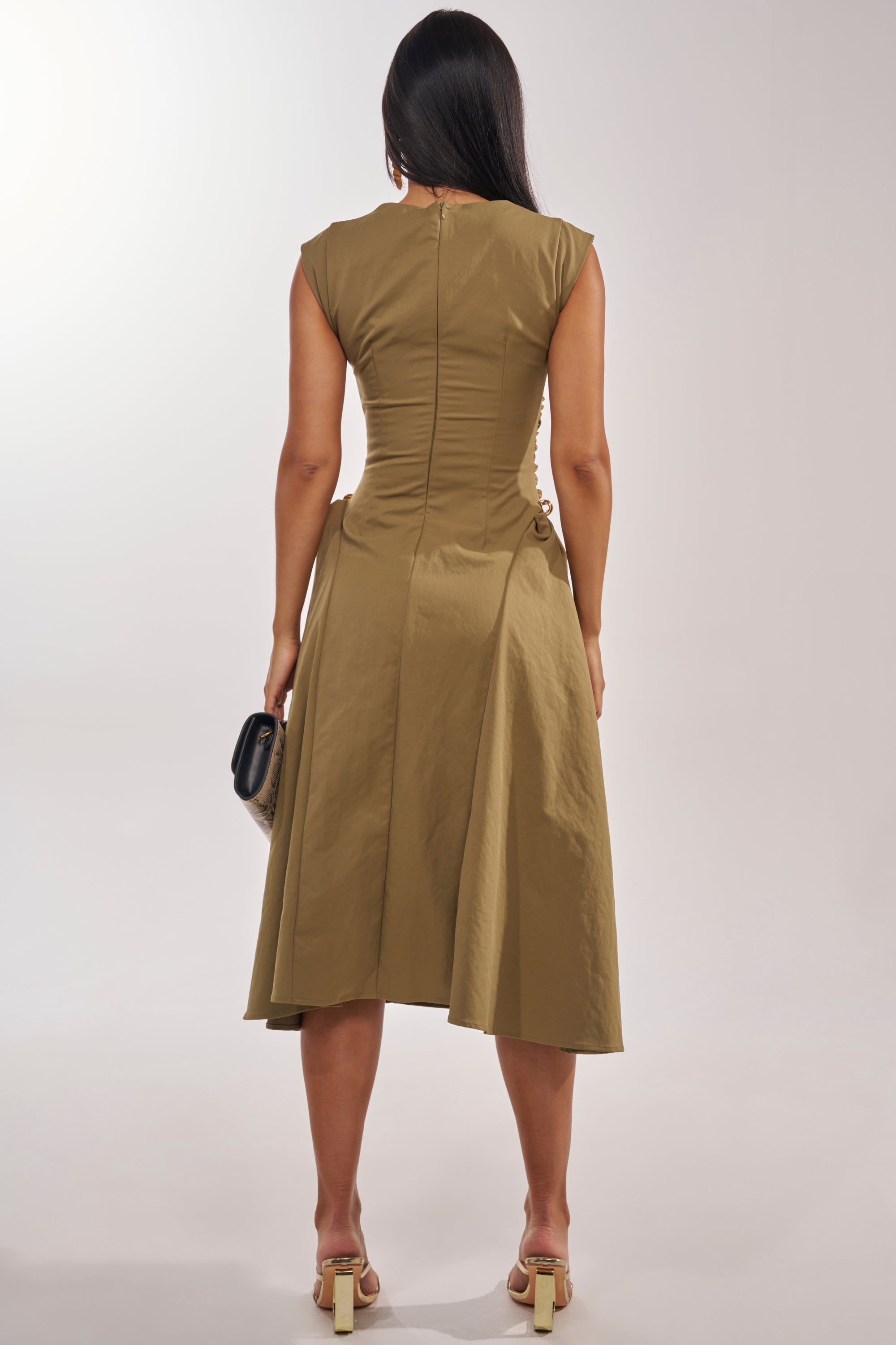 A woman with long dark hair faces away, wearing the IT'S MY TIME MIDI DRESS IN OLIVE with a high neck and cap sleeves. She pairs it with beige heels and a black clutch with gold accents against a plain light background. 5