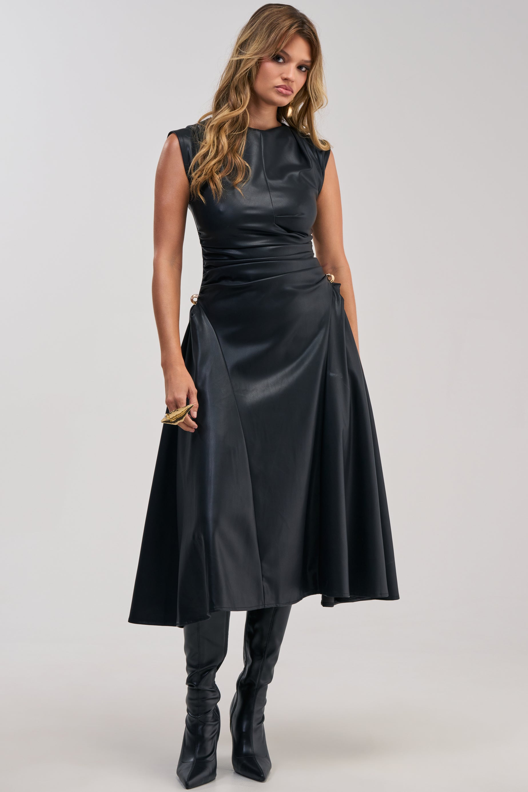 A woman with wavy hair models the IT’S MY TIME MIDI DRESS IN BLACK PU LEATHER, featuring a fitted bodice and flared skirt that make it perfect for cocktail parties. The sleeveless design is styled with tall black boots, as she stands against a plain background.