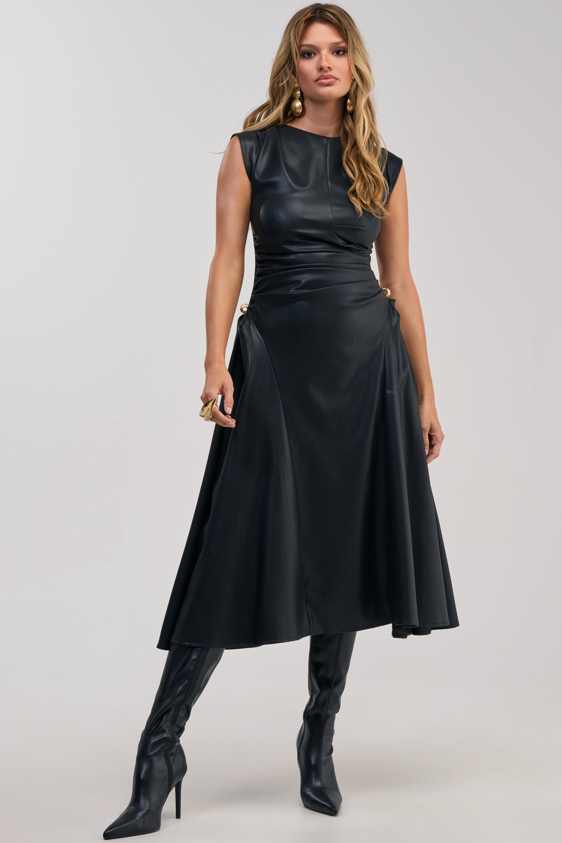 A woman stands confidently against a plain background, wearing the IT’S MY TIME MIDI DRESS IN BLACK PU LEATHER—a sleeveless, flared black faux leather midi dress paired with knee-high heeled boots—ready to make a striking entrance in this modern take on the classic cocktail party look.