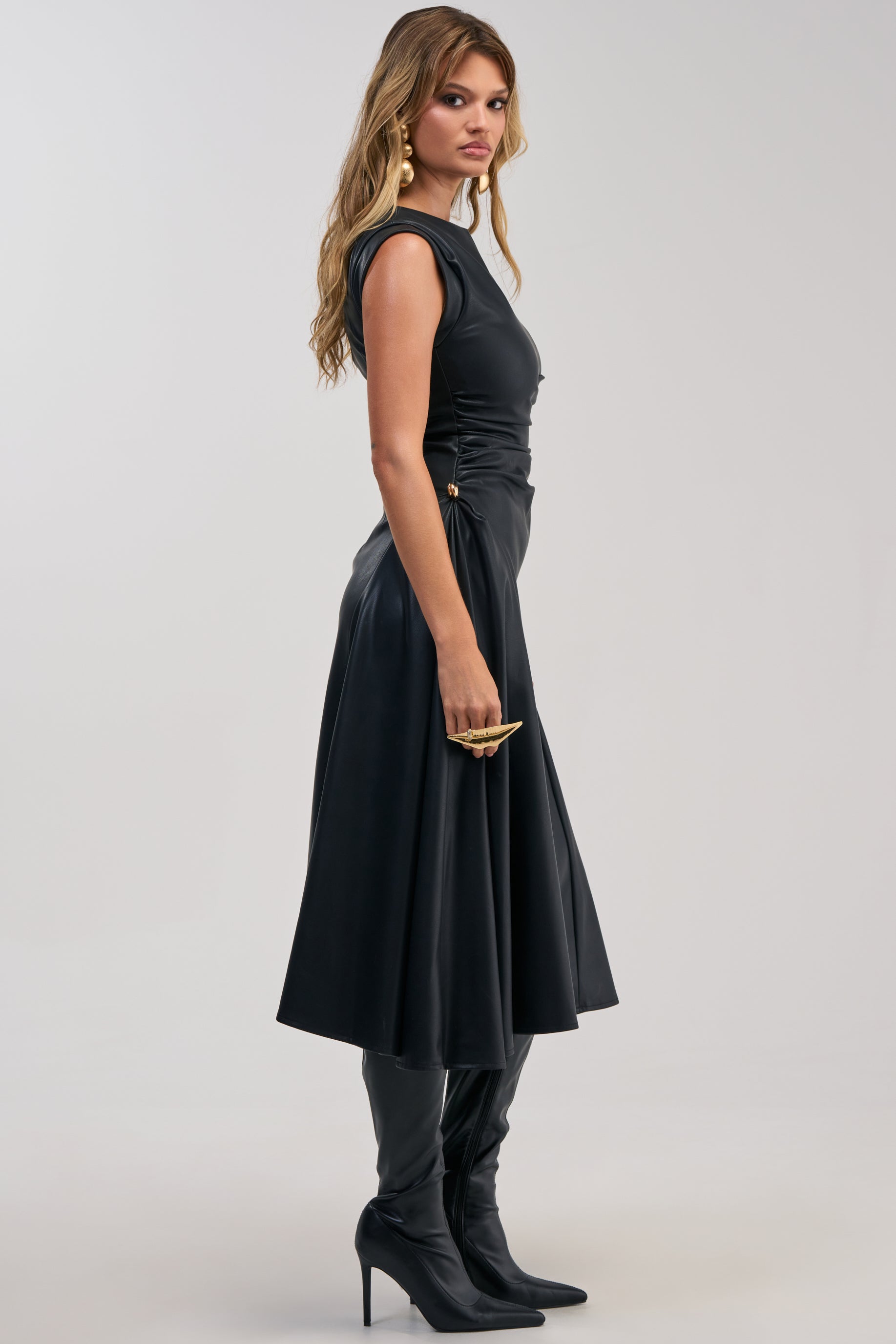 A woman stands sideways against a plain light background, holding a gold clutch and wearing the IT’S MY TIME MIDI DRESS IN BLACK PU LEATHER—a sleeveless, ruched-waist piece that’s elegant for cocktail parties when paired with high-heeled black boots.