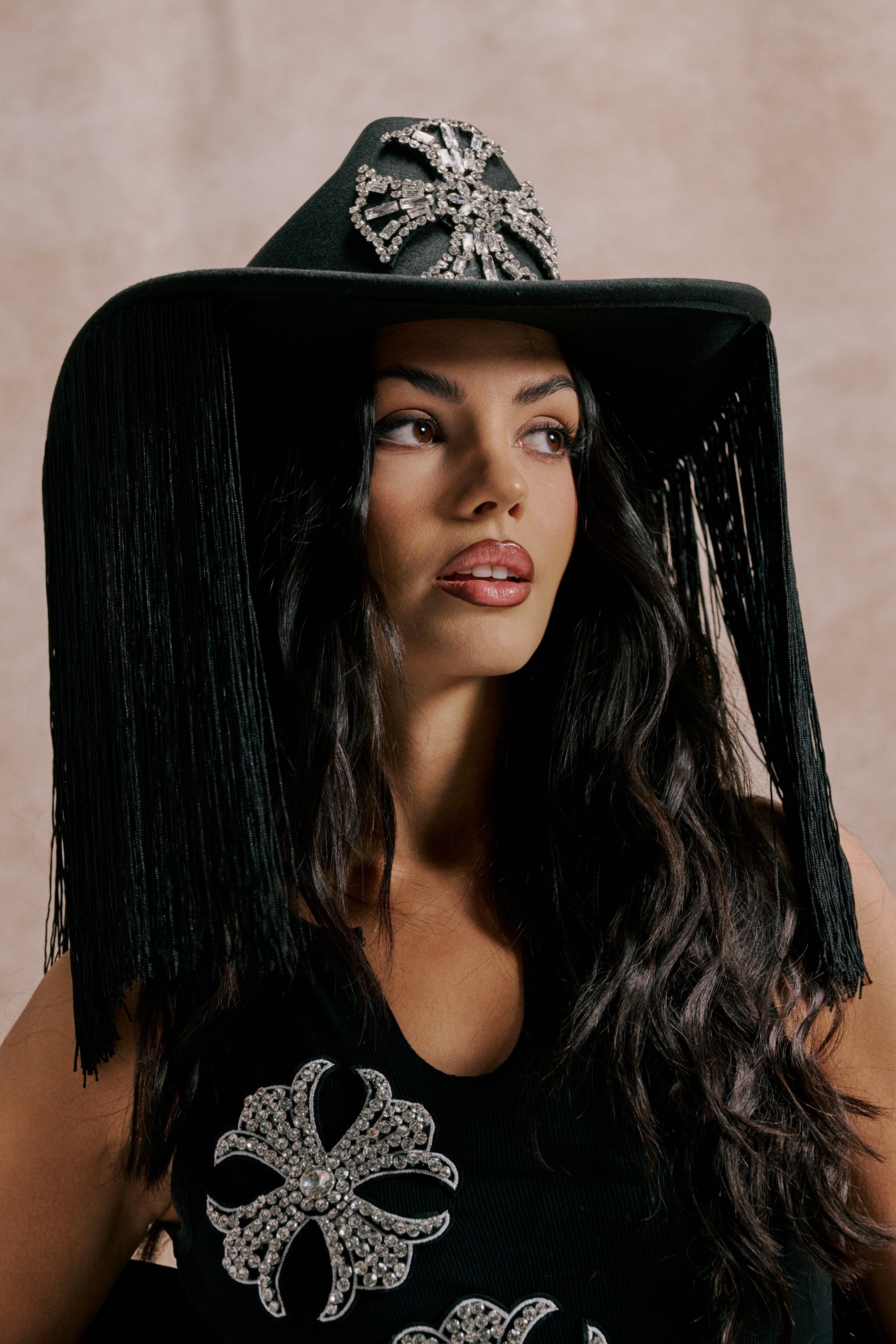 A woman with long dark hair wears the CALL MY HATERS FRINGE BRIM HAT, adorned with silver rhinestones and black fringe, paired with a black top featuring matching rhinestone accents. 0