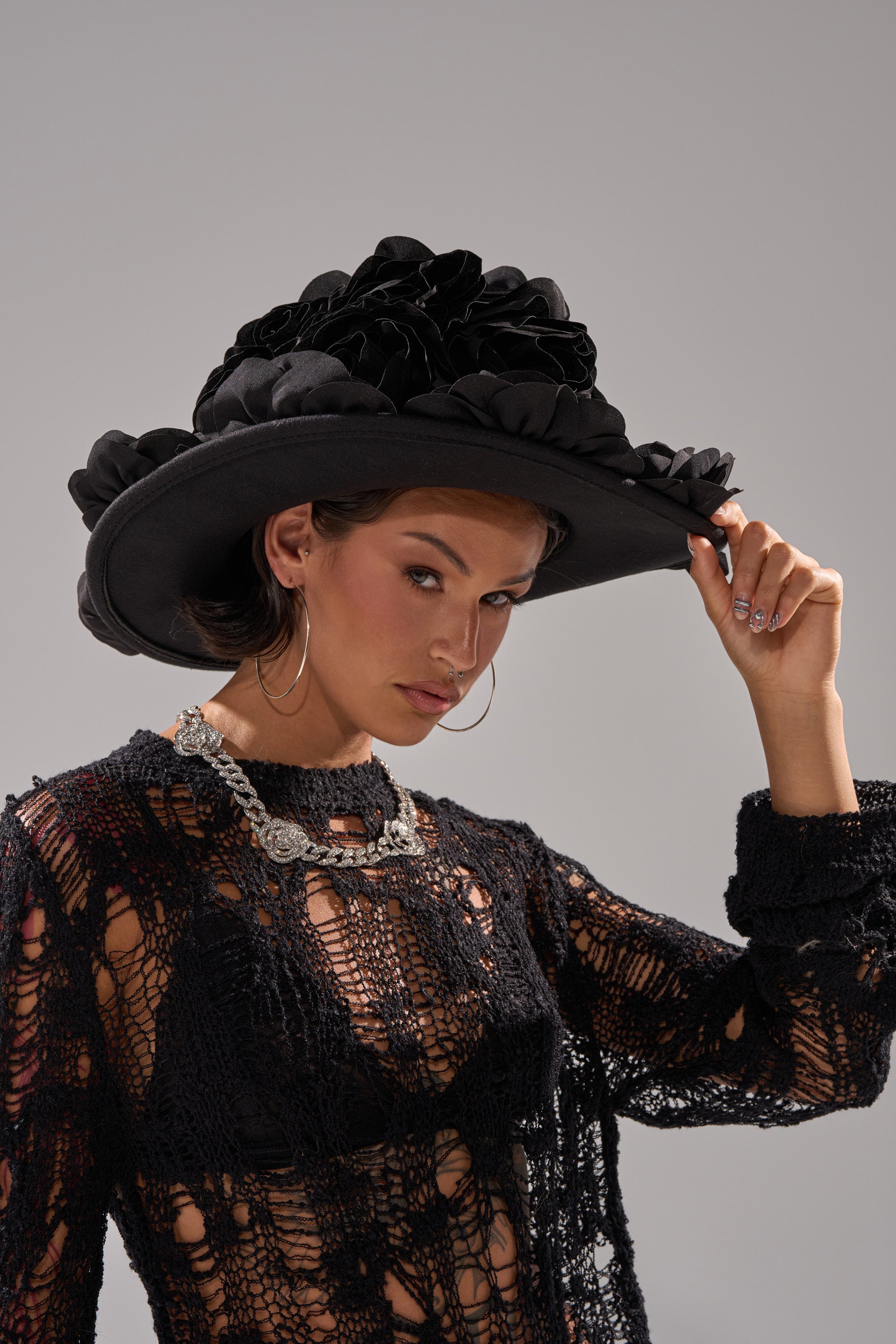 A woman wearing the DARK ROSE FLORAL COWBOY HAT with 3D floral details, a sheer black crochet top, silver hoop earrings, and a chunky chain necklace poses while holding the brim of her hat. 1