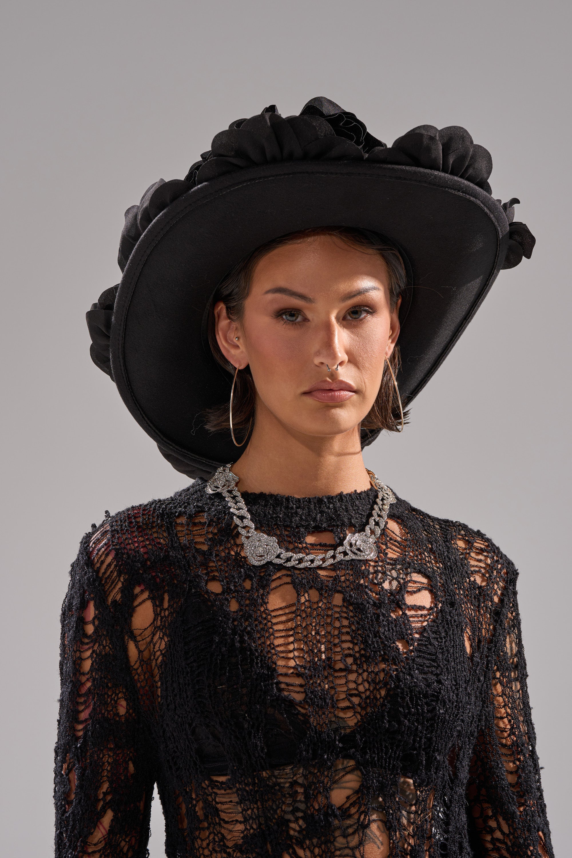 A woman stands against a plain gray background wearing the DARK ROSE FLORAL COWBOY HAT, hoop earrings, a chunky silver necklace, and a black crochet top layered over a black bra. 2