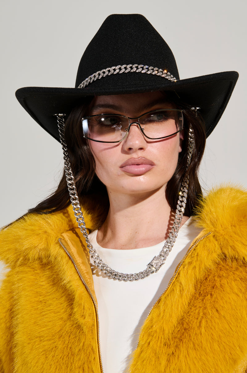 Wearing the LET IT RIDE CHAIN COWBOY HAT, a woman completes her look with aviator sunglasses, a yellow fur coat, and a white top as she gazes forward against a neutral backdrop. 0