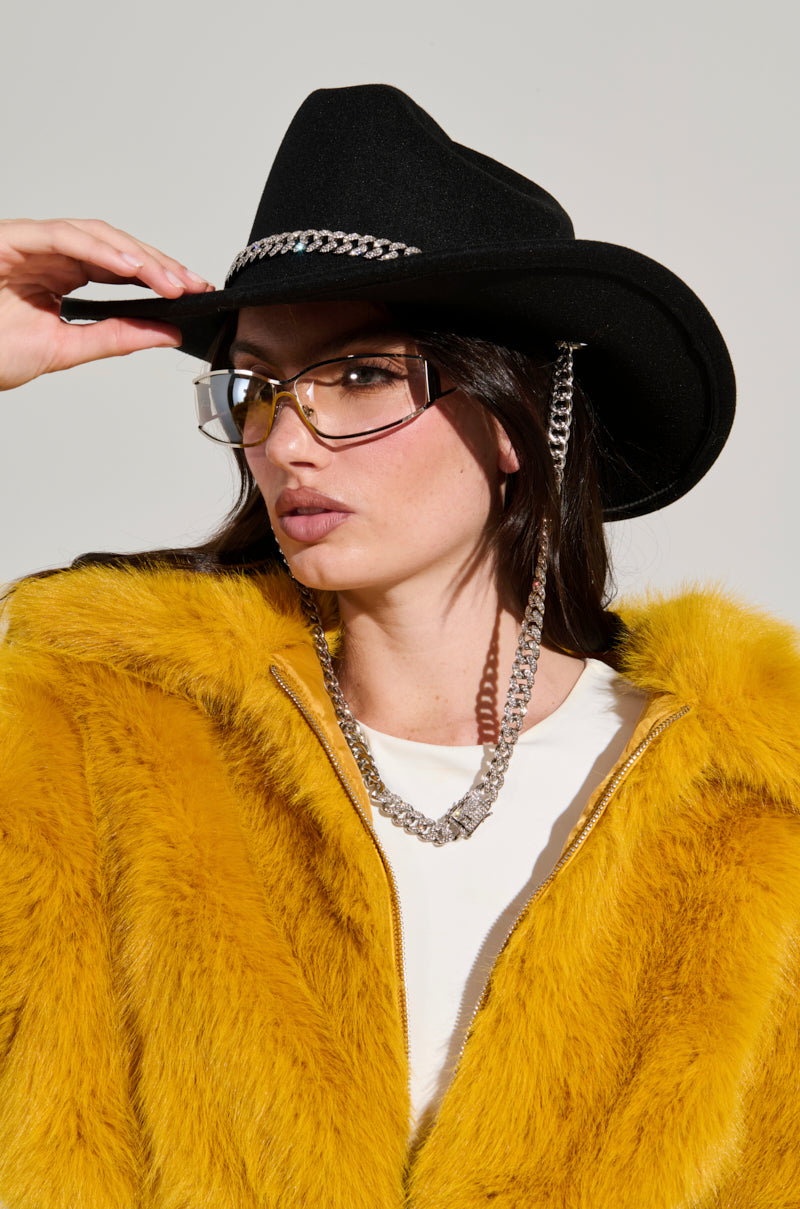 A person wearing the LET IT RIDE CHAIN COWBOY HAT, clear glasses, a rhinestone embellished necklace, and a yellow faux fur jacket poses against a neutral background. They have a serious expression and are slightly tilting the hat. 1