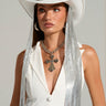 A woman in "THE QUEEN CHAINMAIL COWBOY HAT," a rhinestone embellished white cowboy hat with long silver tassels, wearing a sleeveless outfit adorned with large buttons, hoop earrings, and a chunky chain necklace featuring a cross pendant stands against a plain background.