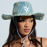 A woman wears the "IN MY CITY COWBOY HAT," a denim-inspired design with "NYC" embroidery and an adjustable chin strap. She pairs it with a black lace top, gazing slightly to the side against a soft backdrop.