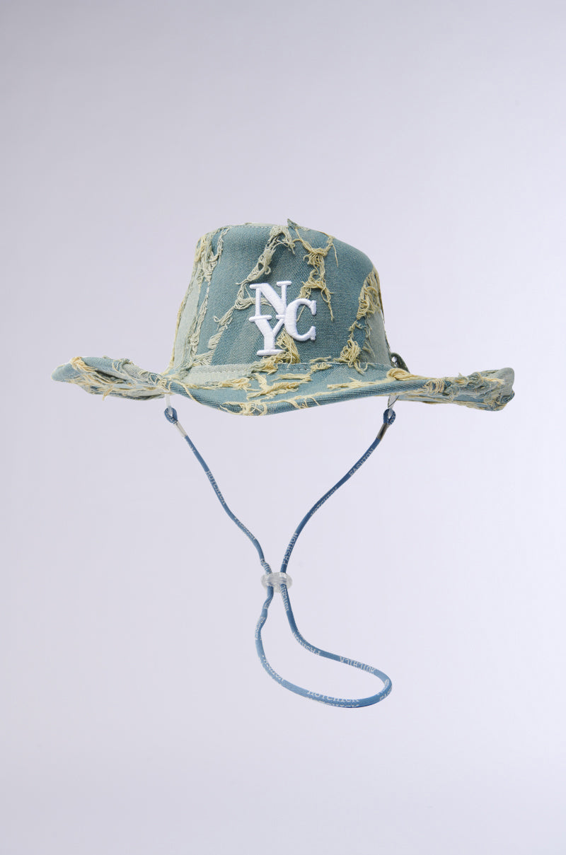 The "IN MY CITY COWBOY HAT" is a denim-inspired distressed light blue hat with frayed edges and yellow accents, featuring "NYC" embroidered in white on the front. It has an adjustable chin strap with a toggle and is set against a plain gradient background.