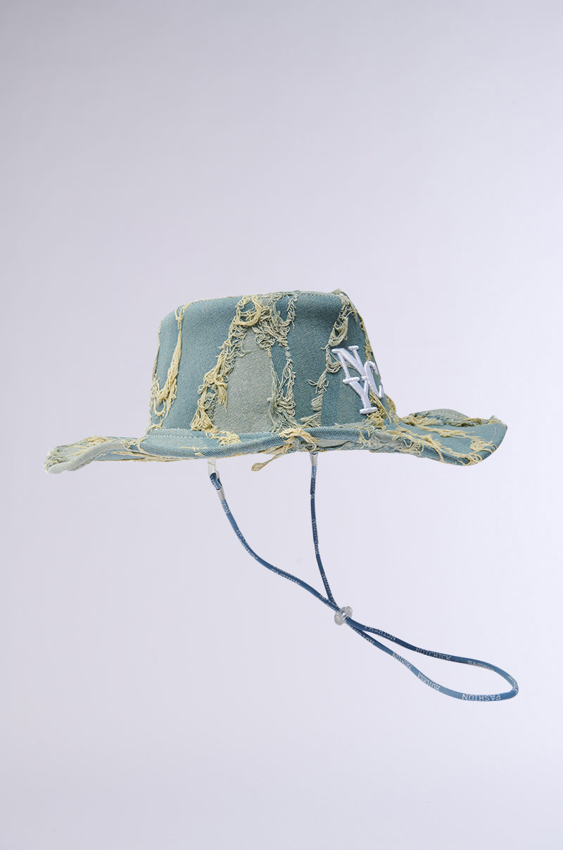 The "IN MY CITY COWBOY HAT" is a distressed denim bucket hat with frayed edges, a wide brim, and an adjustable chin-strap. It features an "NY" logo and NYC embroidery on a plain light background.