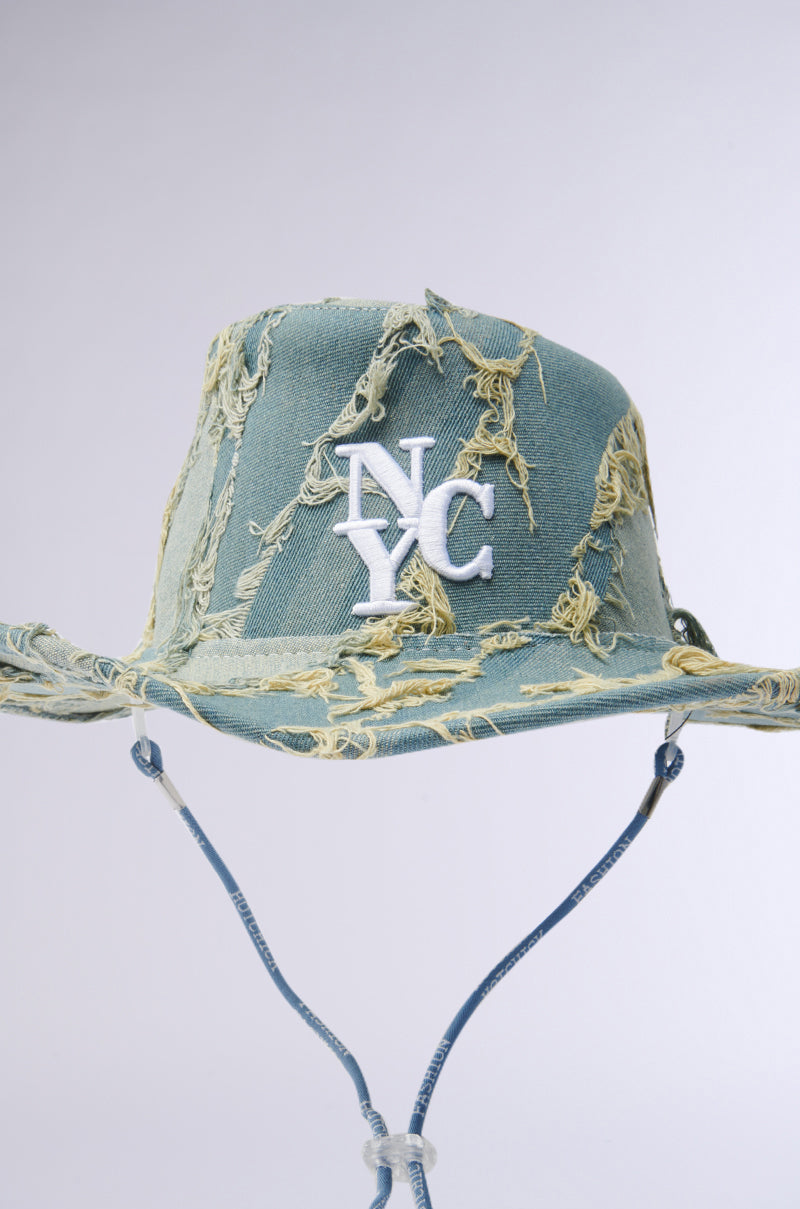 The IN MY CITY COWBOY HAT is a distressed green hat with frayed edges, featuring a white NYC logo. It has an adjustable chin strap and a slightly curved brim, offering a rugged, denim-inspired cowboy hat appearance.