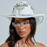 Sporting a BOUJEE PEARL COWBOY HAT adorned with pearls and rhinestones, a woman showcases her long dark hair under stylish sunglasses. A pearl necklace brings elegance, while her white top with pearl accents stands out against a plain light backdrop.