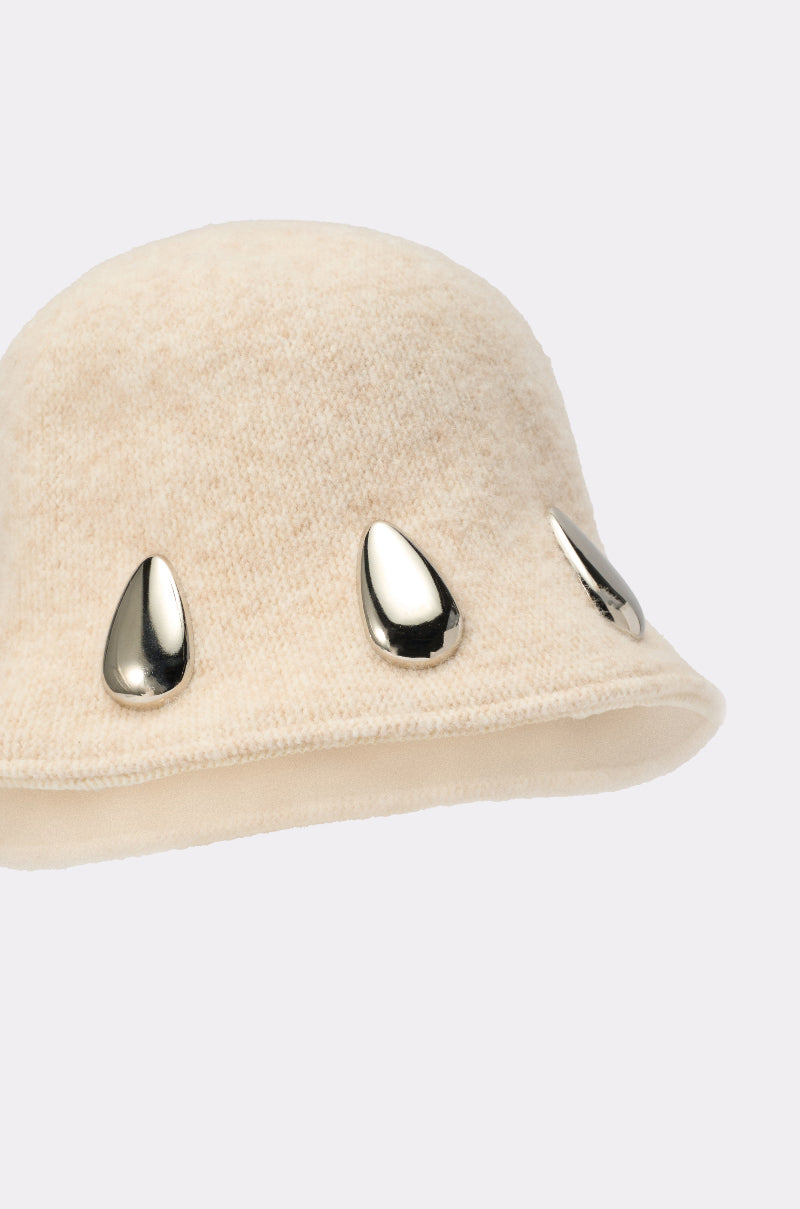 The OUTSIDE METAL DETAIL BUCKET HAT is a beige, slouchy bucket hat with a soft texture and three shiny silver teardrop-shaped metal accents on one side, shown against a plain light background. 4
