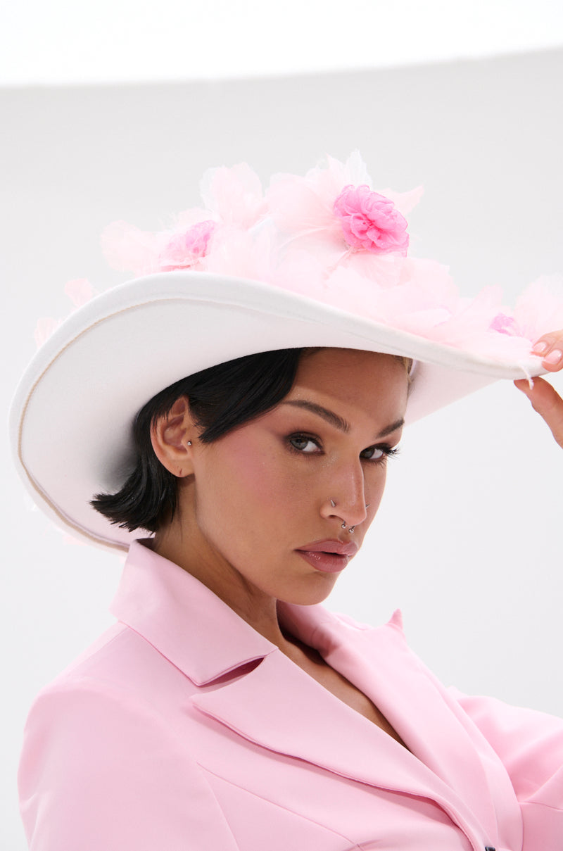 A woman with short dark hair wears a pale pink blazer and the TO THE RACES FLOWER HAT, looking at the camera as she holds the brim of her chic, wide-brimmed accessory adorned with pink flowers.