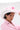 A woman with short dark hair wears a pale pink blazer and the TO THE RACES FLOWER HAT, looking at the camera as she holds the brim of her chic, wide-brimmed accessory adorned with pink flowers.