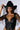 A person wearing the "I SEE WHAT I NEED COWBOY HAT" featuring rhinestone detailing and a netted veil, along with a black leather corset top and a tattoo on their arm, looks directly at the camera. One hand is holding the brim of the hat, and they have a serious expression.