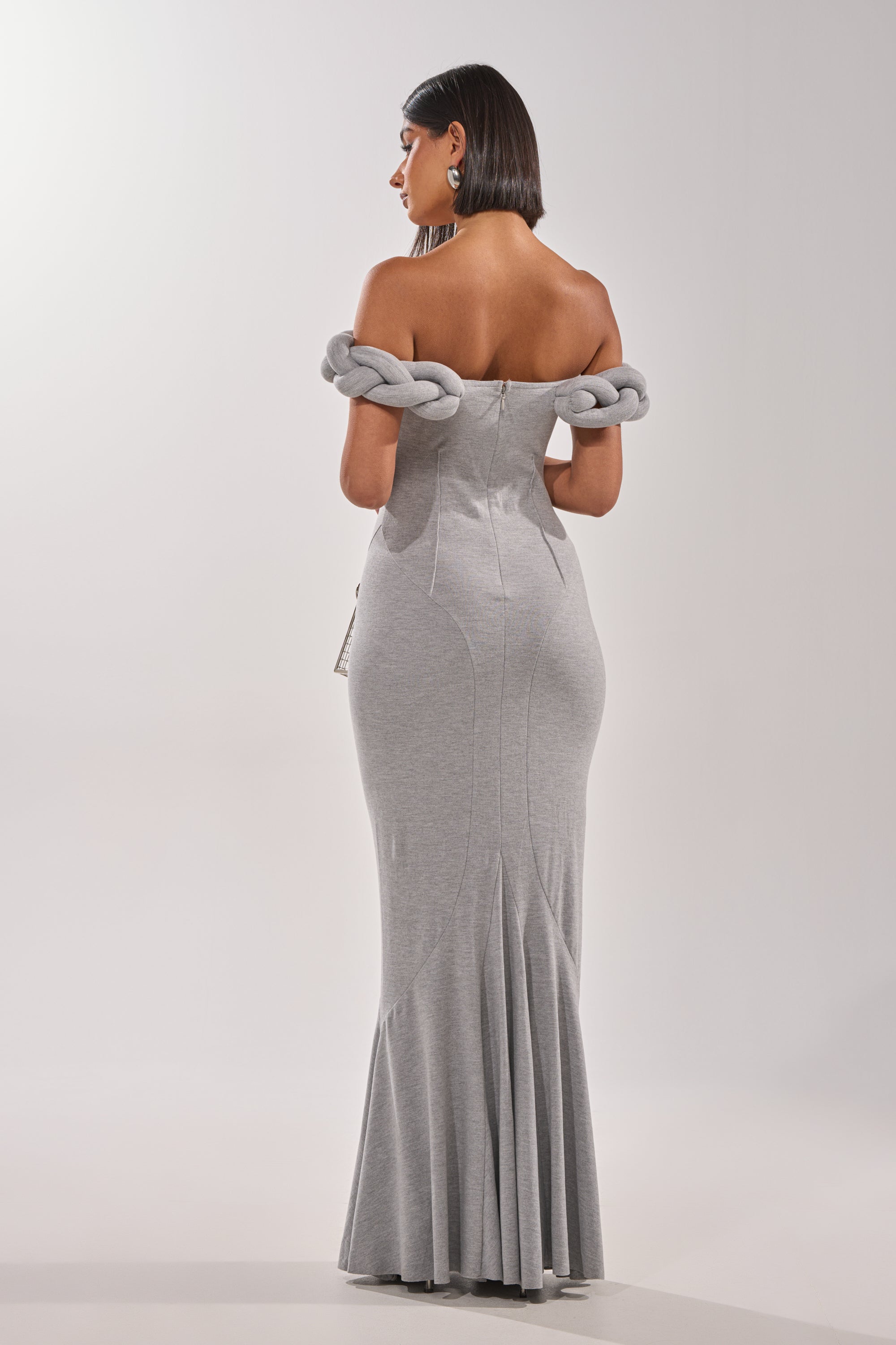 A woman faces away in the GODDESS HOUR KNIT MAXI DRESS—a grey, off-the-shoulder formal gown with braided knit sleeves. The fitted maxi flares at the hem. She has dark, straight hair and stands against a plain white background. 4