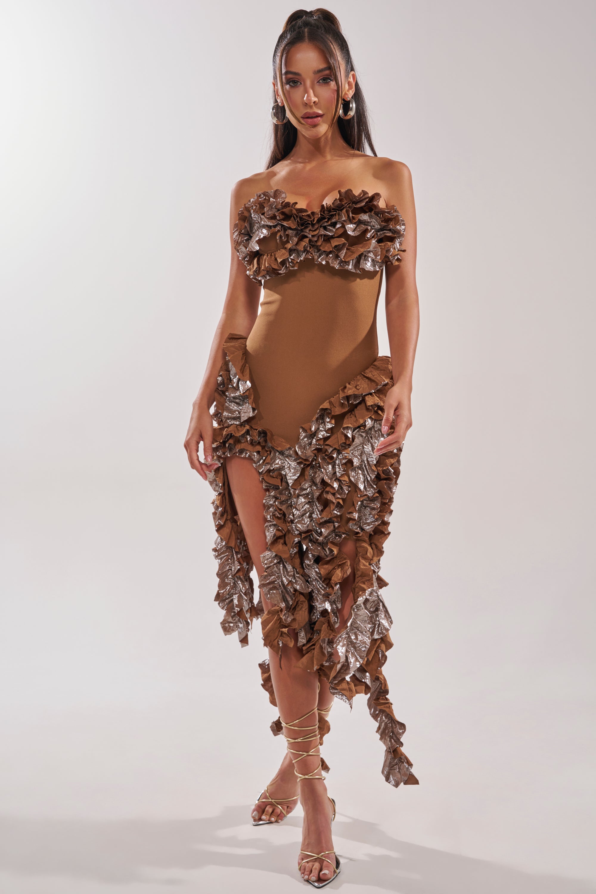 A woman poses confidently in the CITY LIGHTS RUFFLE MIDI DRESS IN BRONZE, featuring a strapless sweetheart neckline and ruffled asymmetrical hem, styled with strappy heels against a simple light backdrop. 1