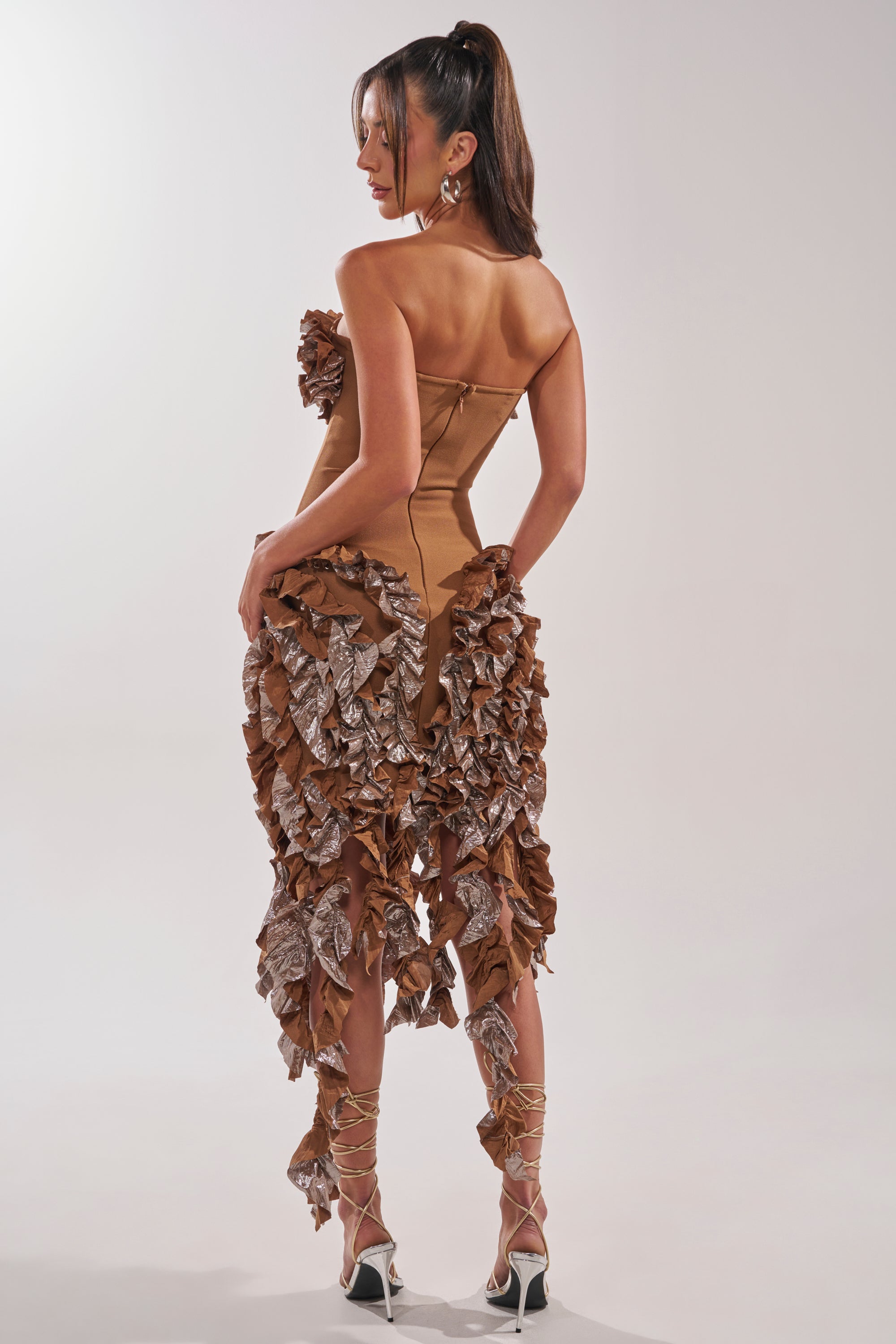 A woman wears the CITY LIGHTS RUFFLE MIDI DRESS IN BRONZE—a strapless, sweetheart neckline dress with ruffle trim. She stands in profile, her long hair in a ponytail, paired with strappy high-heeled sandals. 5