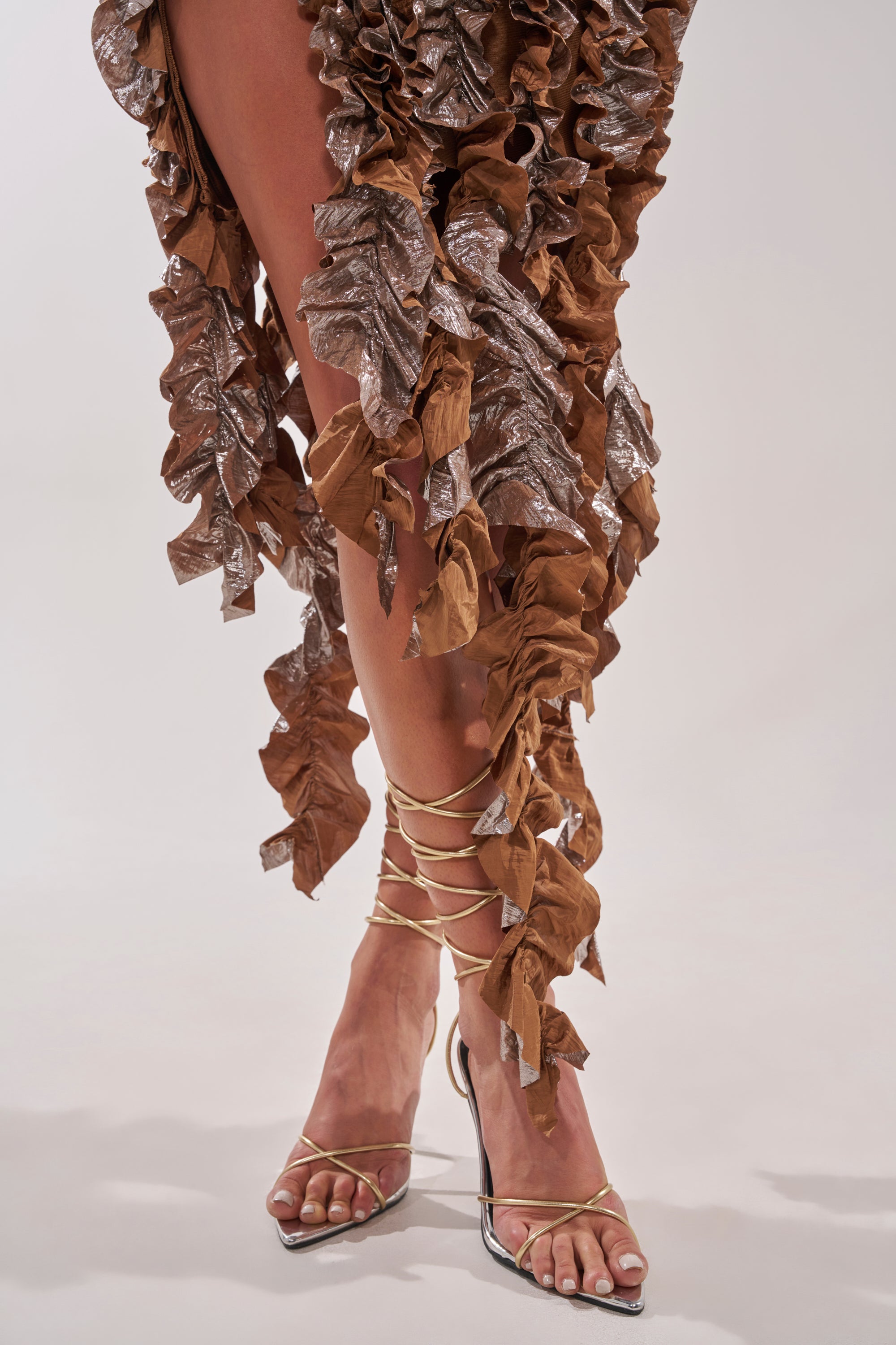 A close-up of a person’s legs wearing the CITY LIGHTS RUFFLE MIDI DRESS IN BRONZE and beige lace-up high-heeled sandals, standing on a plain light background. 7