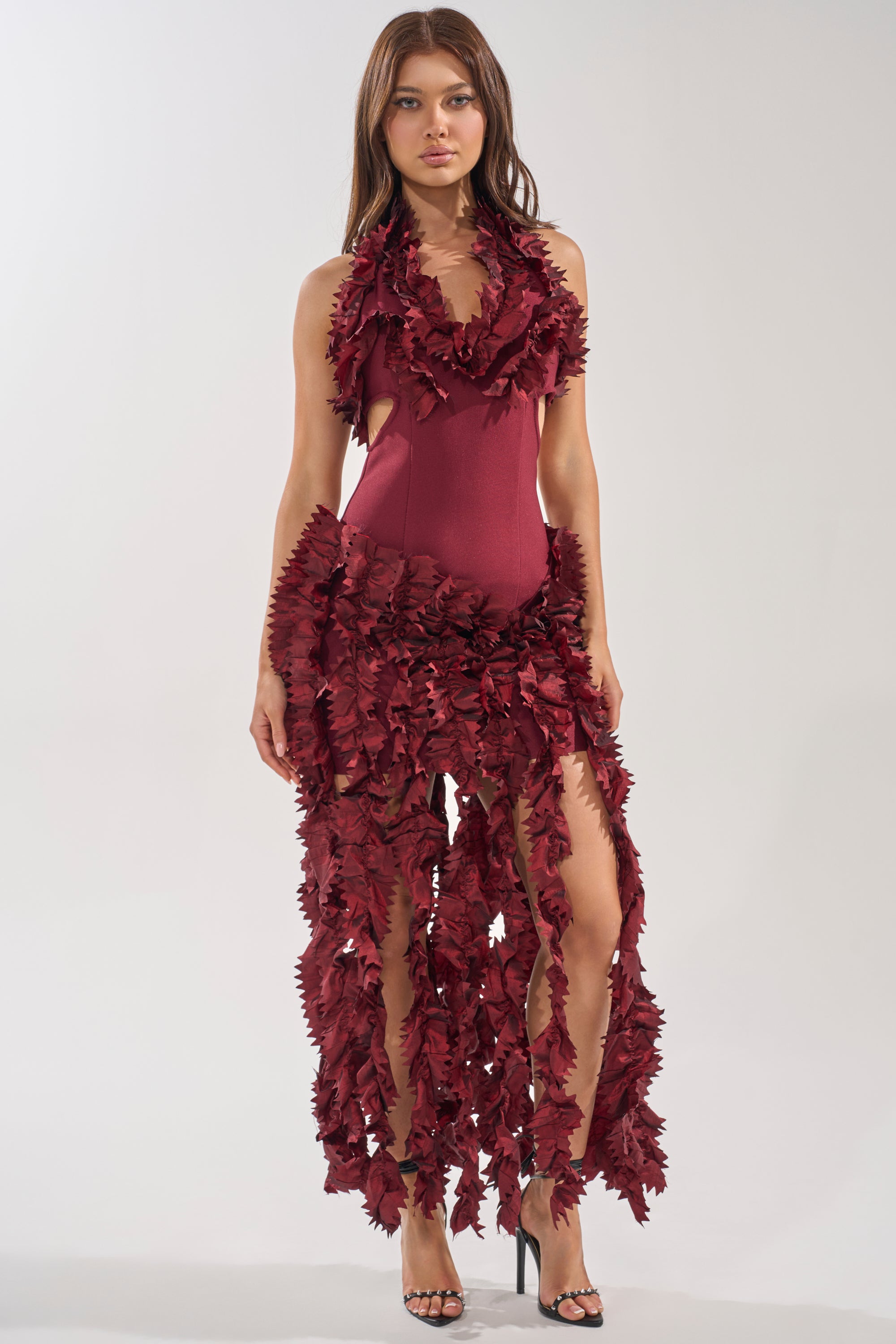 A woman poses in the BEST DRESSED FLOCKED MAXI DRESS, a maroon halter maxi with bold 3D ruffle appliqué from neck to ankle. Paired with black high-heeled sandals against a plain backdrop, it makes an elegant statement for evening occasions.