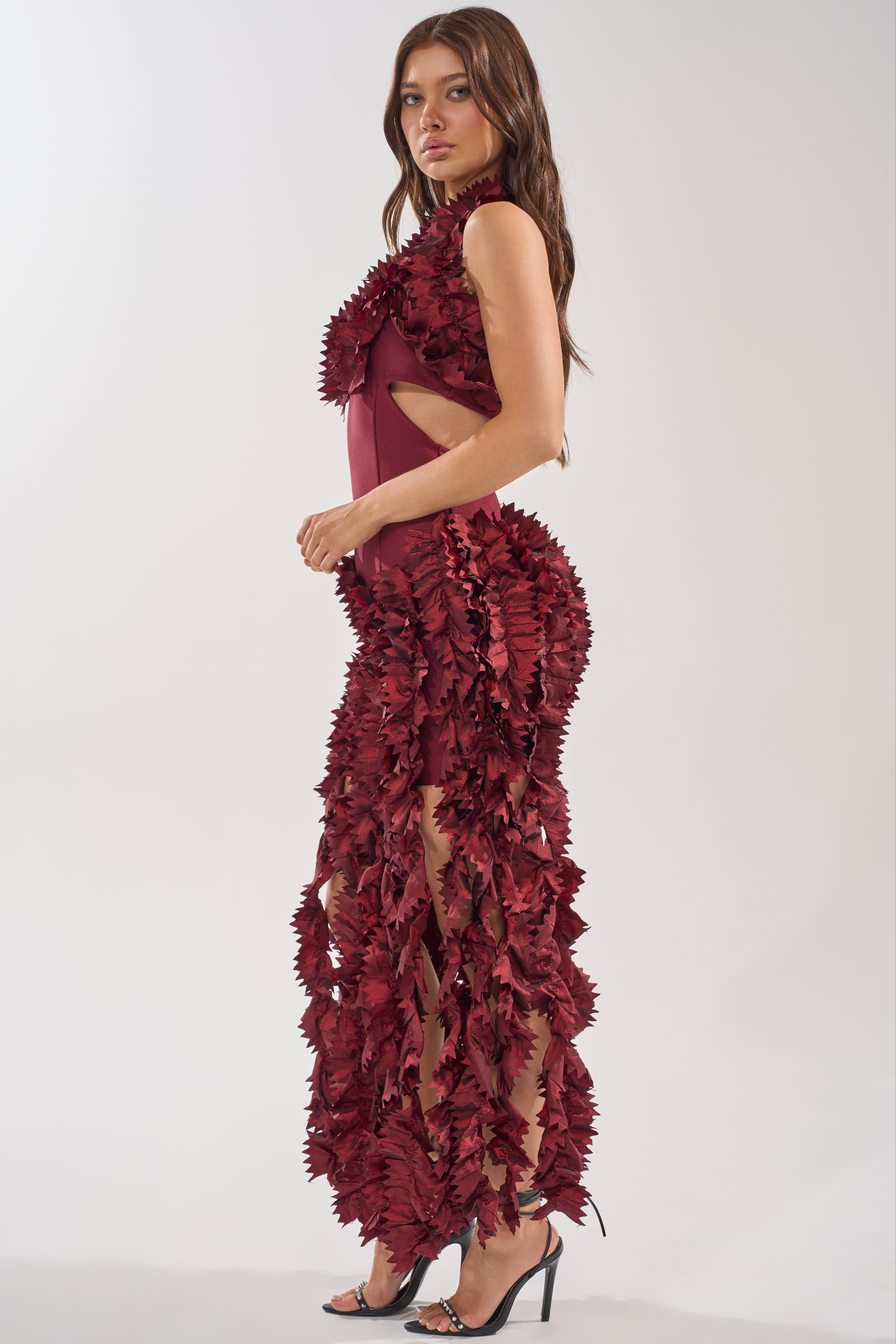 A woman stands in profile wearing the BEST DRESSED FLOCKED MAXI DRESS, featuring a maroon halter style with 3D leaf appliqués, side cut-outs, and sheer sections. She pairs it with black heels—ideal for an elegant evening look.