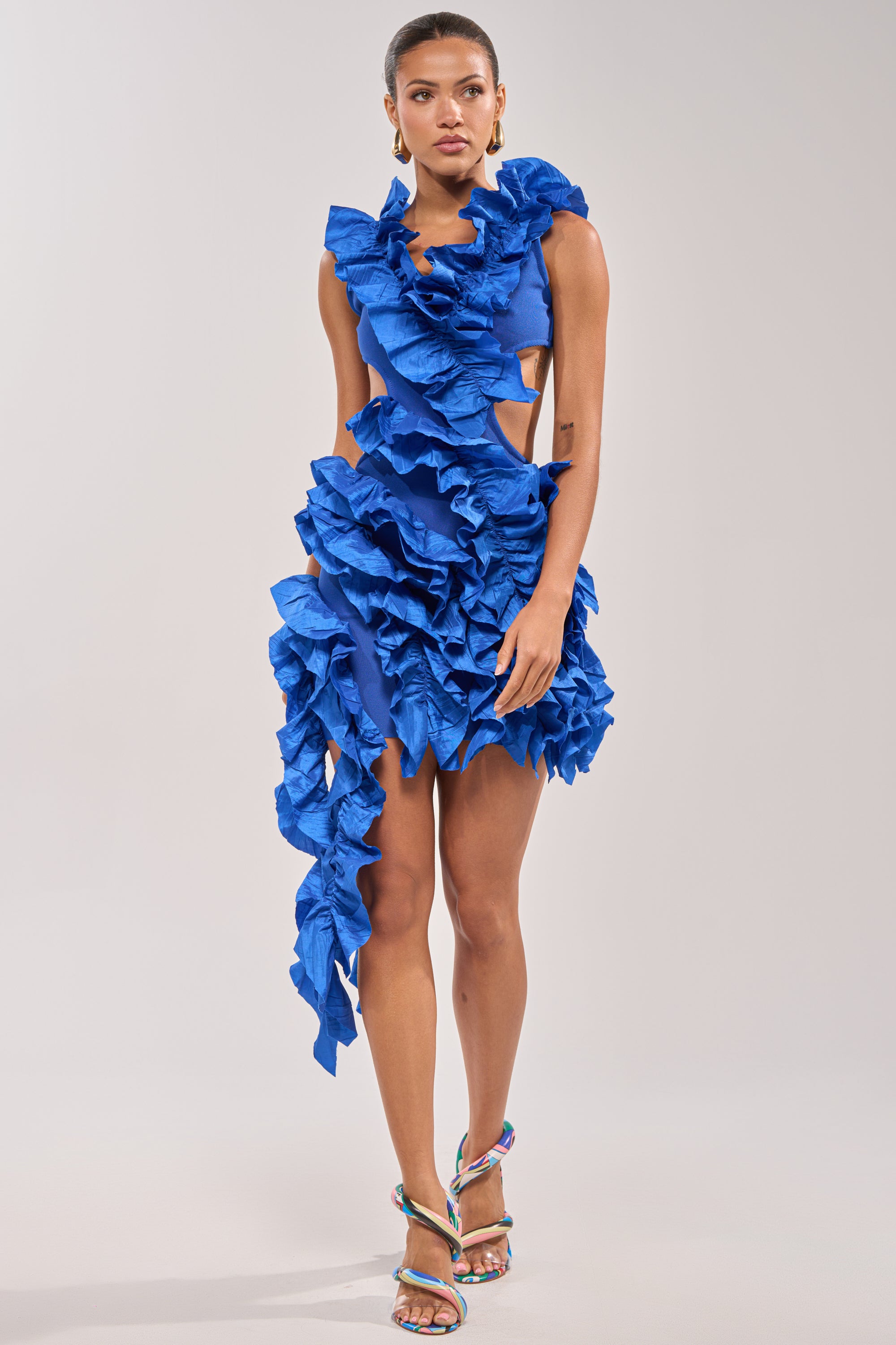 A woman wears the CRAZIER THAN YOUR EX FLOCKED MINI DRESS IN BLUE, featuring vibrant cutouts, ruffle layers, and an asymmetrical hem. She pairs it with colorful strappy heels and gold hoop earrings—ideal for formal occasions.