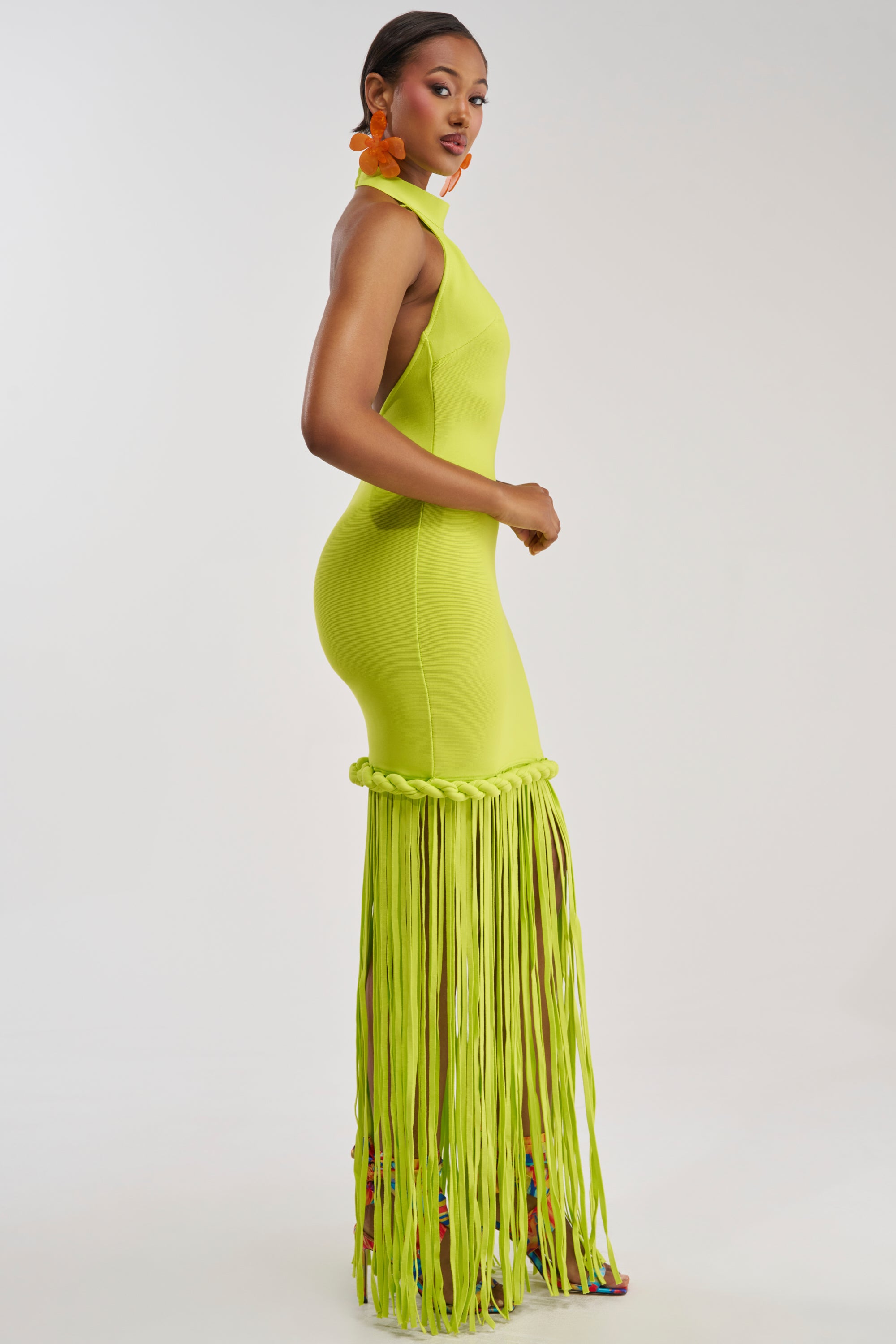 A woman wearing the BEACH THEN CLUB MAXI DRESS IN LIME—a bright lime green, form-fitting maxi dress with long fringe at the bottom—pairs it with orange earrings and colorful shoes while posing in profile against a plain white background.