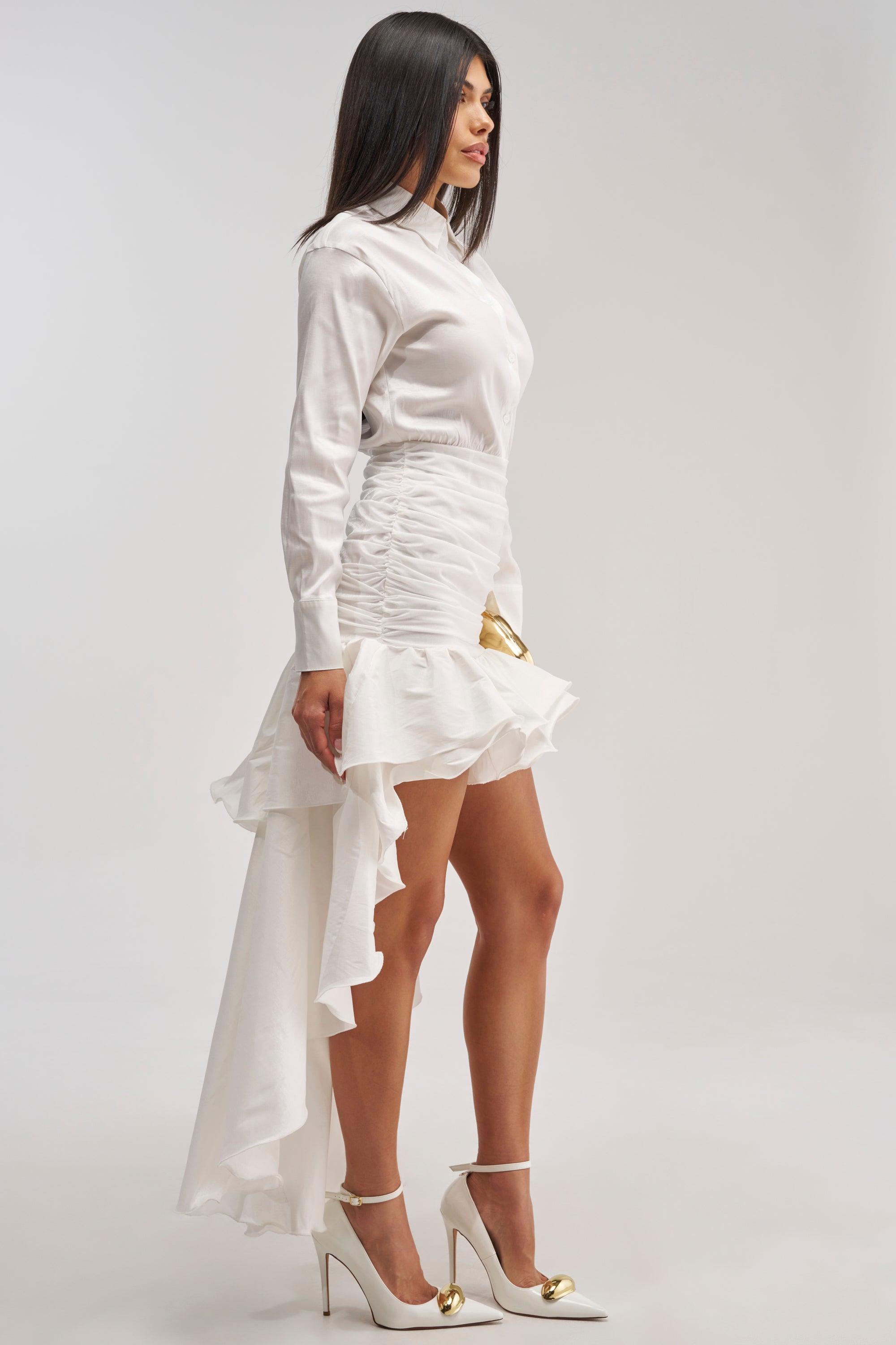 A woman stands in profile against a plain light background, wearing the BACK TO WERK Poplin Maxi Dress in white, featuring a high-low ruffled skirt, styled with white high heels adorned with gold tips.