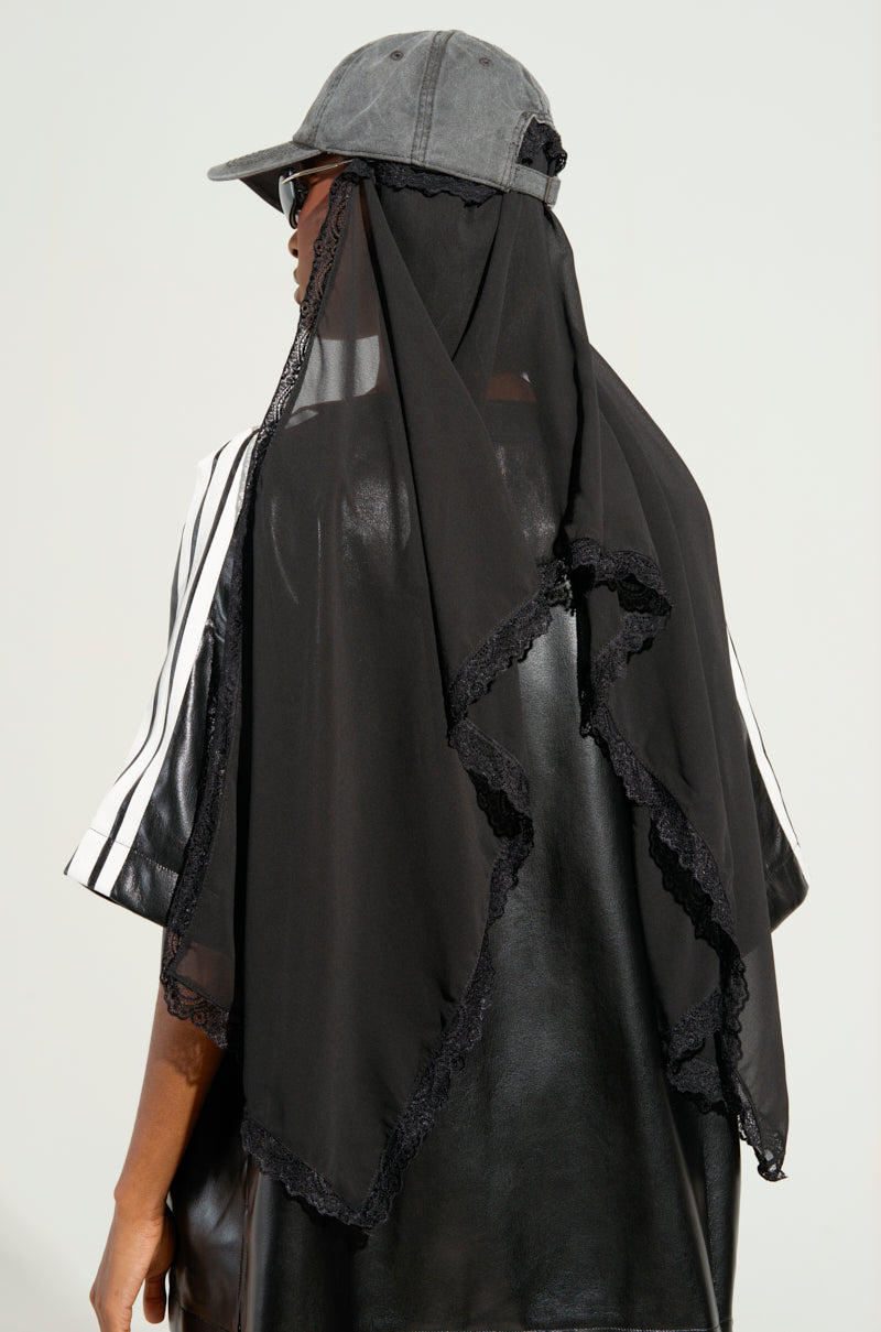 A person wearing the TRINA VEIL SNAPBACK, paired with black sunglasses and a black lace-trimmed headscarf over a shiny black garment with white-striped sleeves, stands facing away from the camera against a plain background. 1