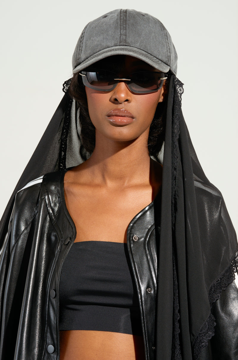 A person wearing a black leather jacket, black tube top, and a TRINA VEIL SNAPBACK cap poses stylishly with sunglasses against a plain background. 2