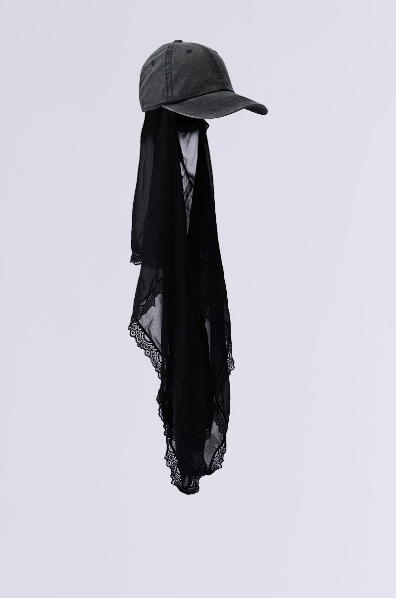 The TRINA VEIL SNAPBACK is a charcoal gray cap with a sleek mesh veil, complemented by a long black lace scarf trailing from the back, all set against a plain light background. 4